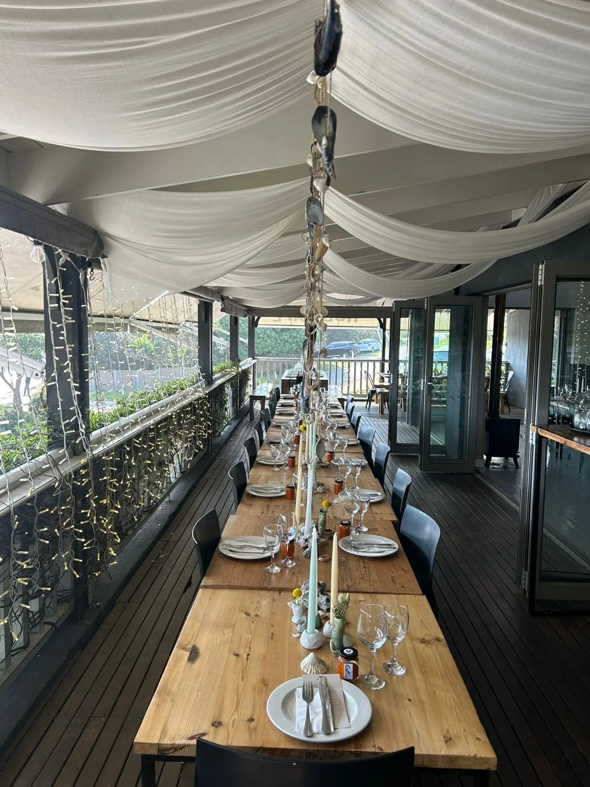 Long dining table set for a meal, with plates, glasses, and utensils, decorated with candles and small decorative items, on a deck with string lights and large white drapes overhead.