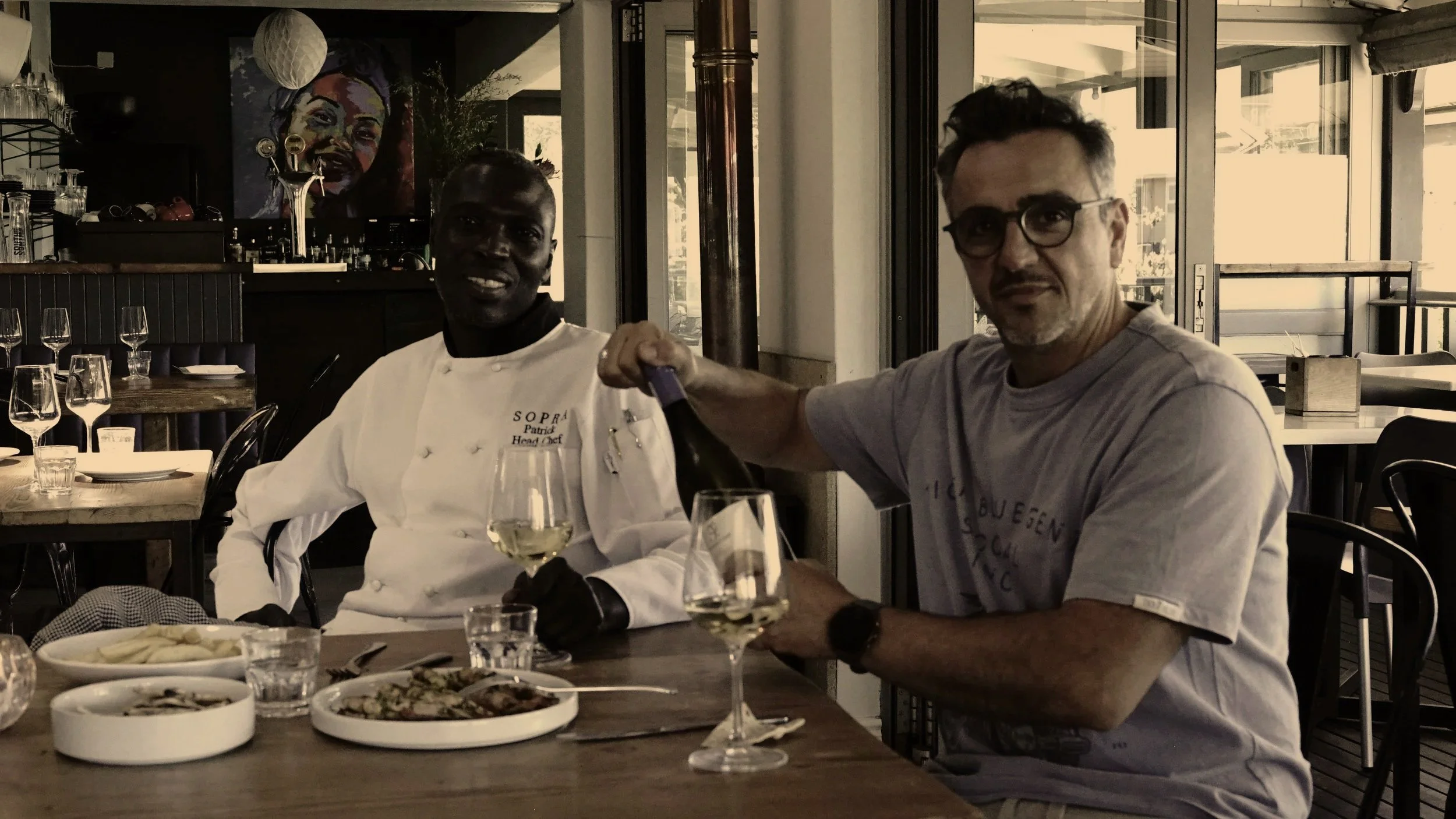 A man in a gray T-shirt pouring white wine into a glass at a restaurant table, with a chef in a white coat smiling beside him. The table has plates of food, wine glasses, and water glasses, in a well-lit dining area with large windows.