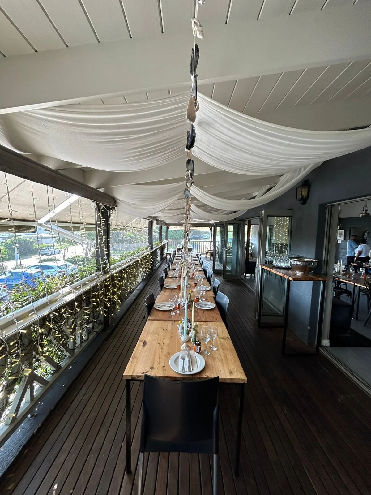 Long outdoor dining table set with plates, glasses, and candles, decorated for an event, under white draped fabric ceiling on a balcony with string lights.