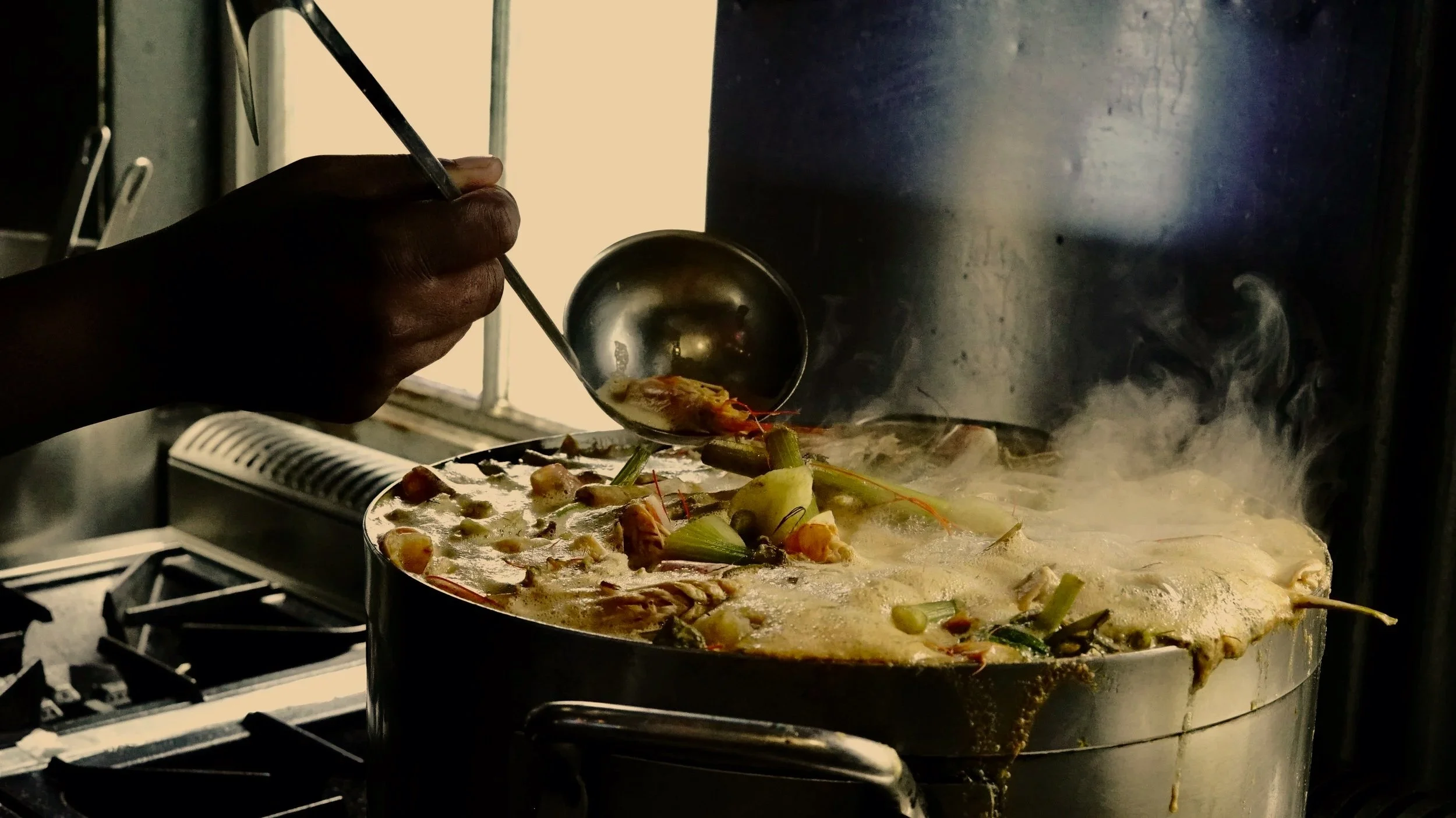 A person serving food from a steaming pot in a kitchen.