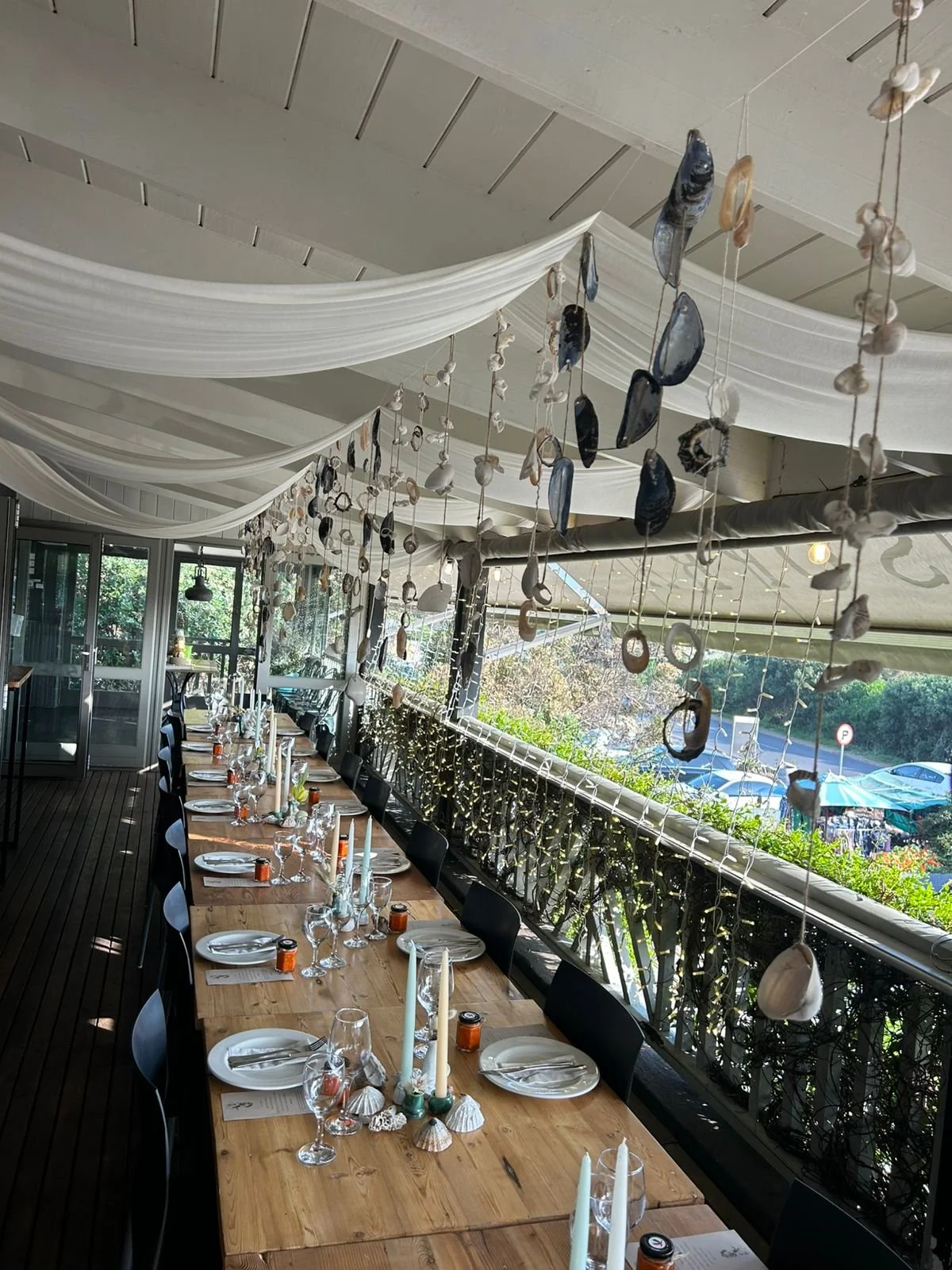 A long dining table set for a meal on a balcony or porch, decorated with candles, seashells, and small orange jars, with hanging seashells and starfish decorations above and ceiling drapes, overlooking a sunny outdoor area.