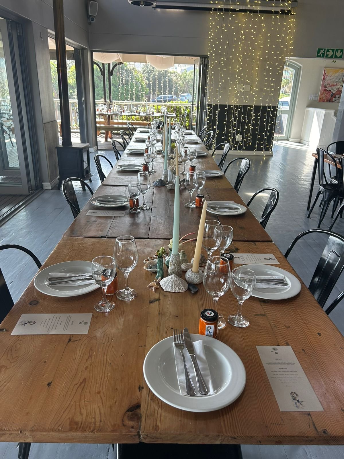 Long wooden dining table set for a meal with plates, silverware, wine glasses, and small decorative shells and candles in the center. Fairy lights hang from the ceiling. Sunny outdoor scenery is visible through open sliding glass doors.