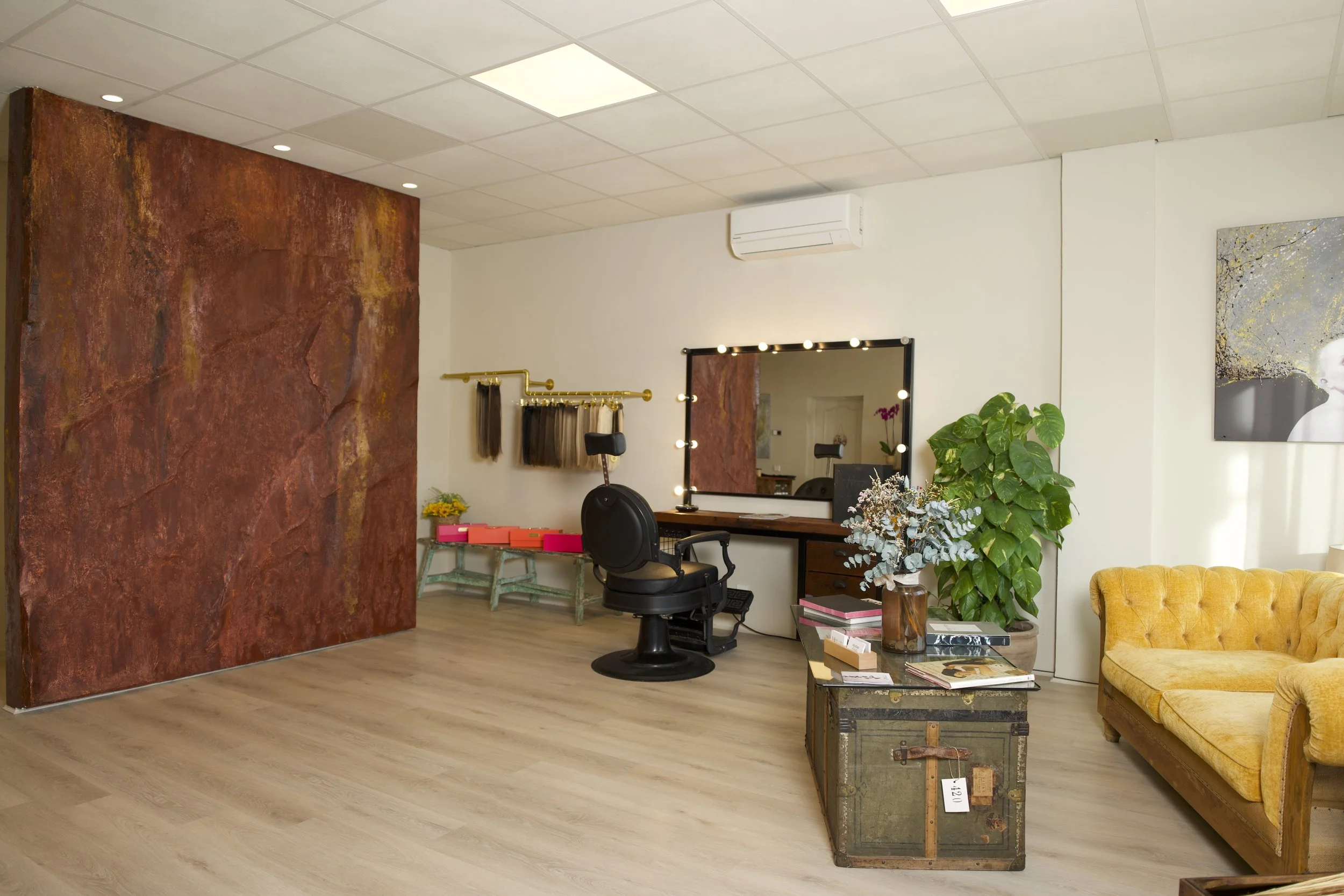 Sala de peluquería con espejo con luces, silla de estilista, maniquí con cabello, sofá mostaza, planta grande, mesa con libros, y pared de color terracota.