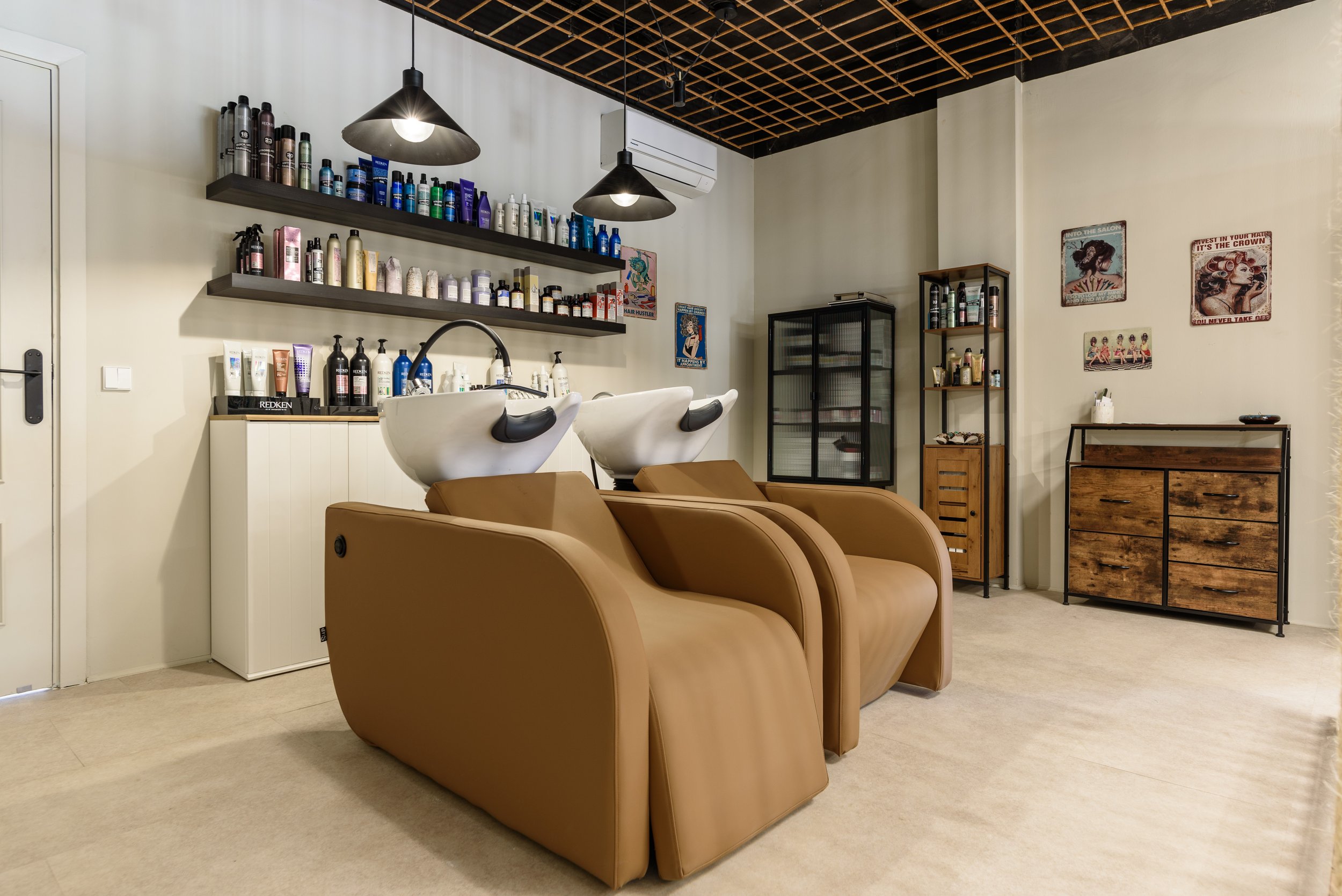 Sala de cabinas de lavado de cabello con sillones de cuero beige y lavabos blancos, estantes con productos para el cabello, muebles de madera y paredes decoradas con posters vintage.