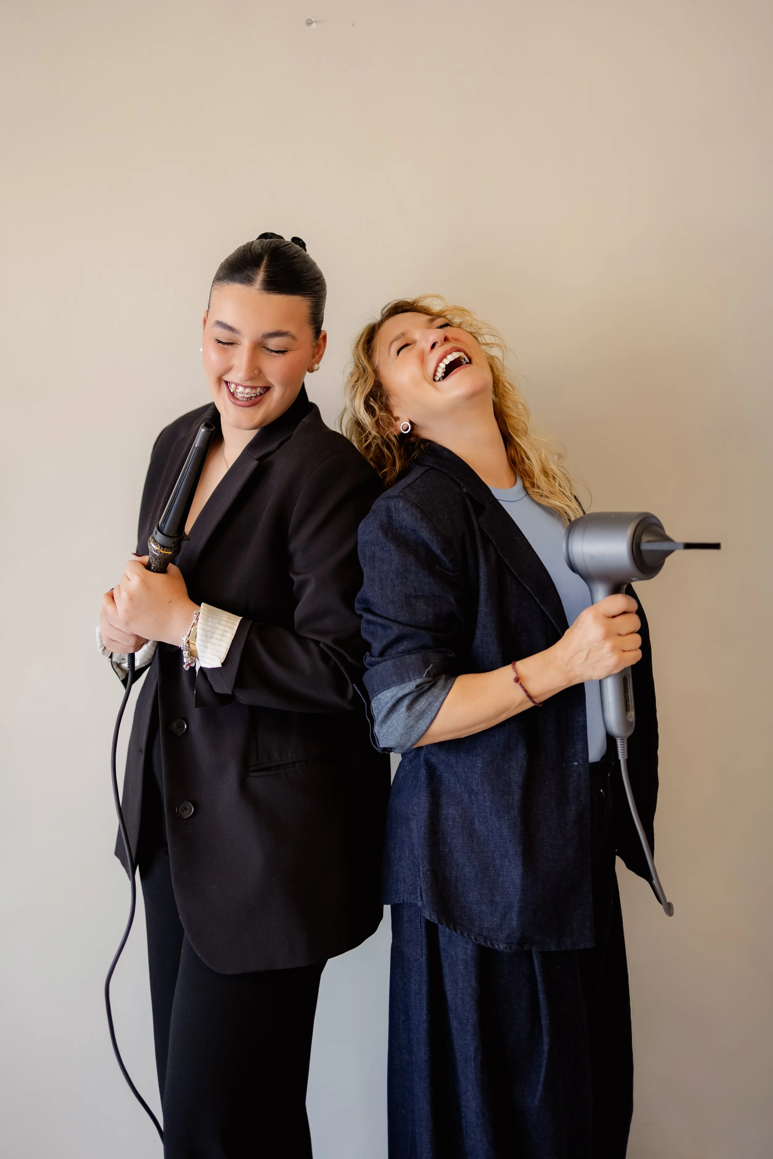 Dos mujeres con cabello recogido, sonriendo y sosteniendo secadores de cabello, una con ropa formal y la otra con ropa casual, de espaldas y apoyadas entre sí.