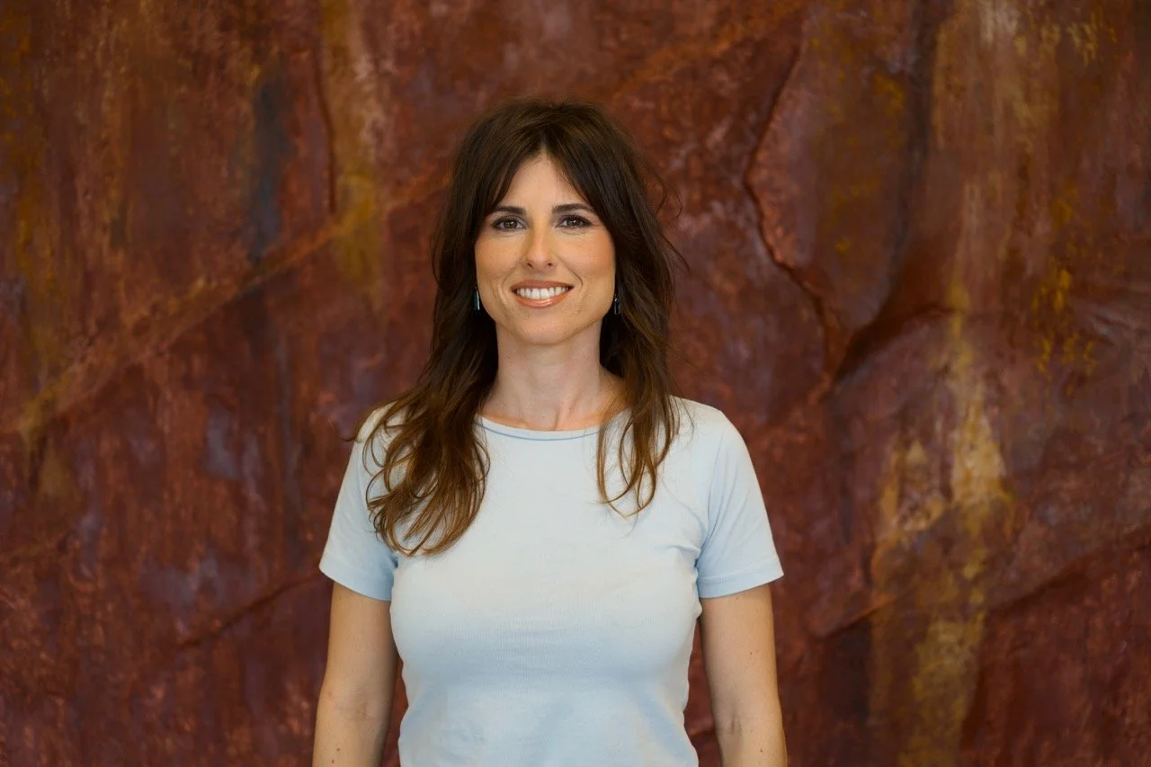 Mujer con cabello castaño y largo, sonriendo, lleva camiseta blanca, fondo de roca rojiza.