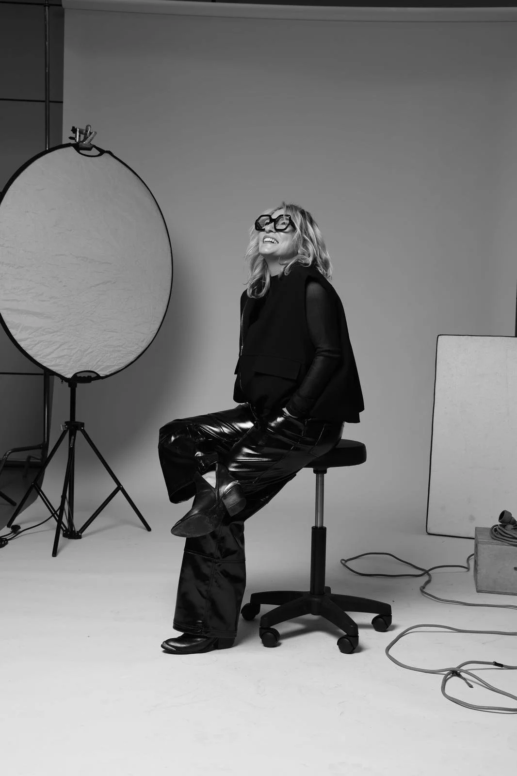 Mujer rubia con gafas grandes, mostrando una sonrisa, en un estudio fotográfico en blanco y negro, sentada en una silla con ruedas, con equipo de iluminación y accesorios alrededor.