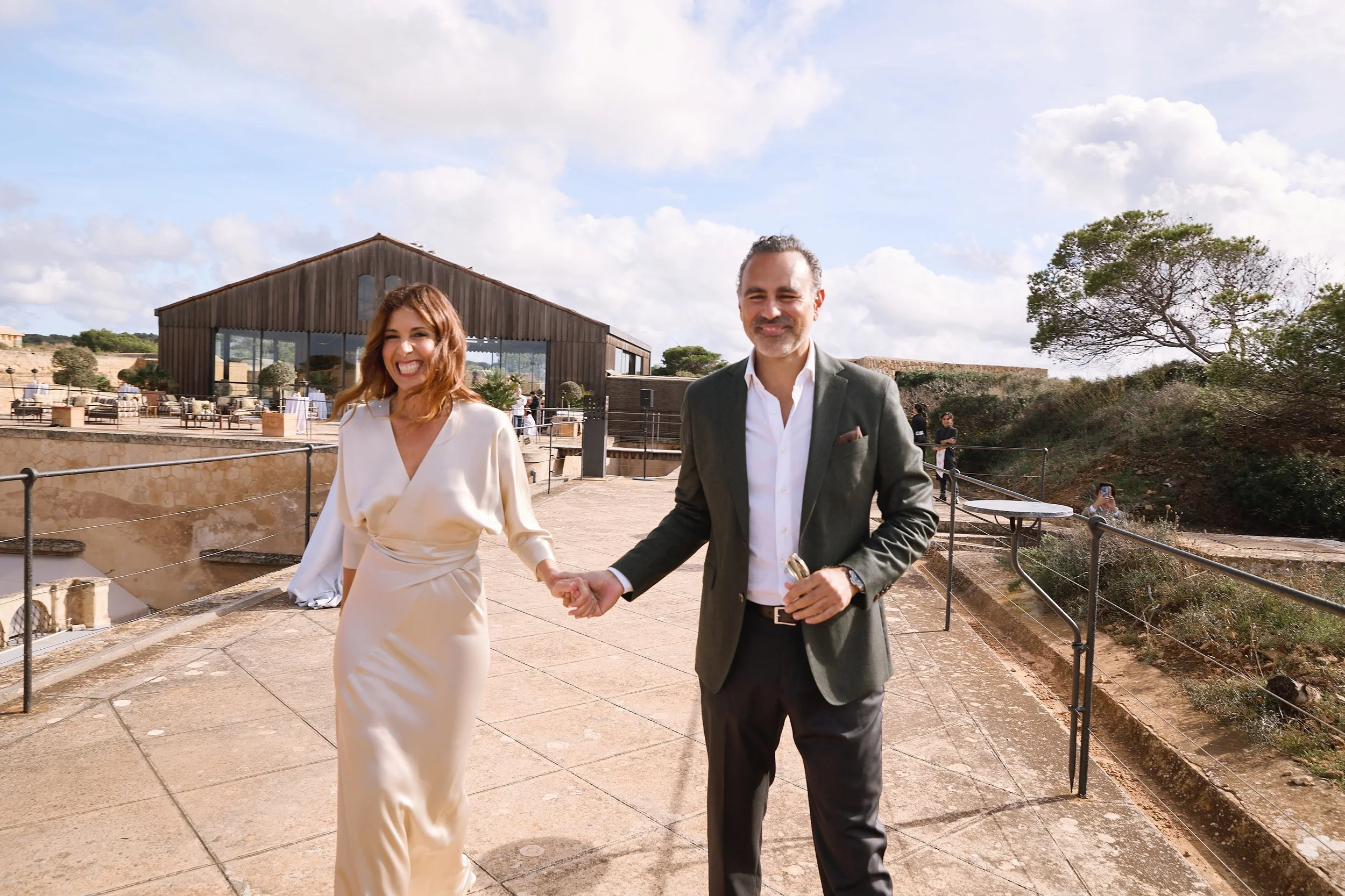 Pareja sonriendo y caminando juntos en un espacio abierto con edificio de madera y naturaleza, durante un día soleado.