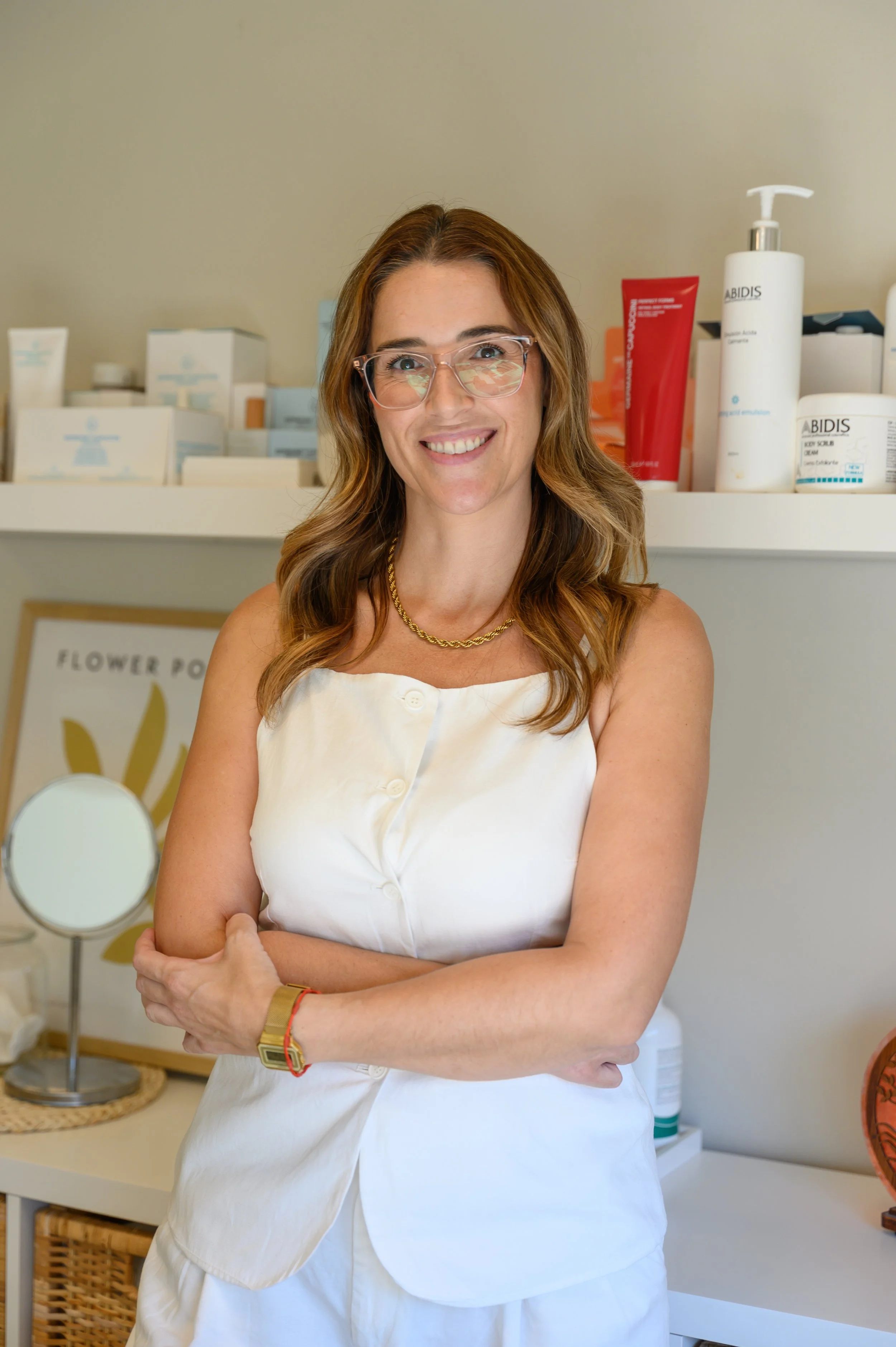 Mujer sonriendo con lentes y ropa blanca, de pie en una tienda de cuidado de la piel, con productos y un espejo en el fondo.