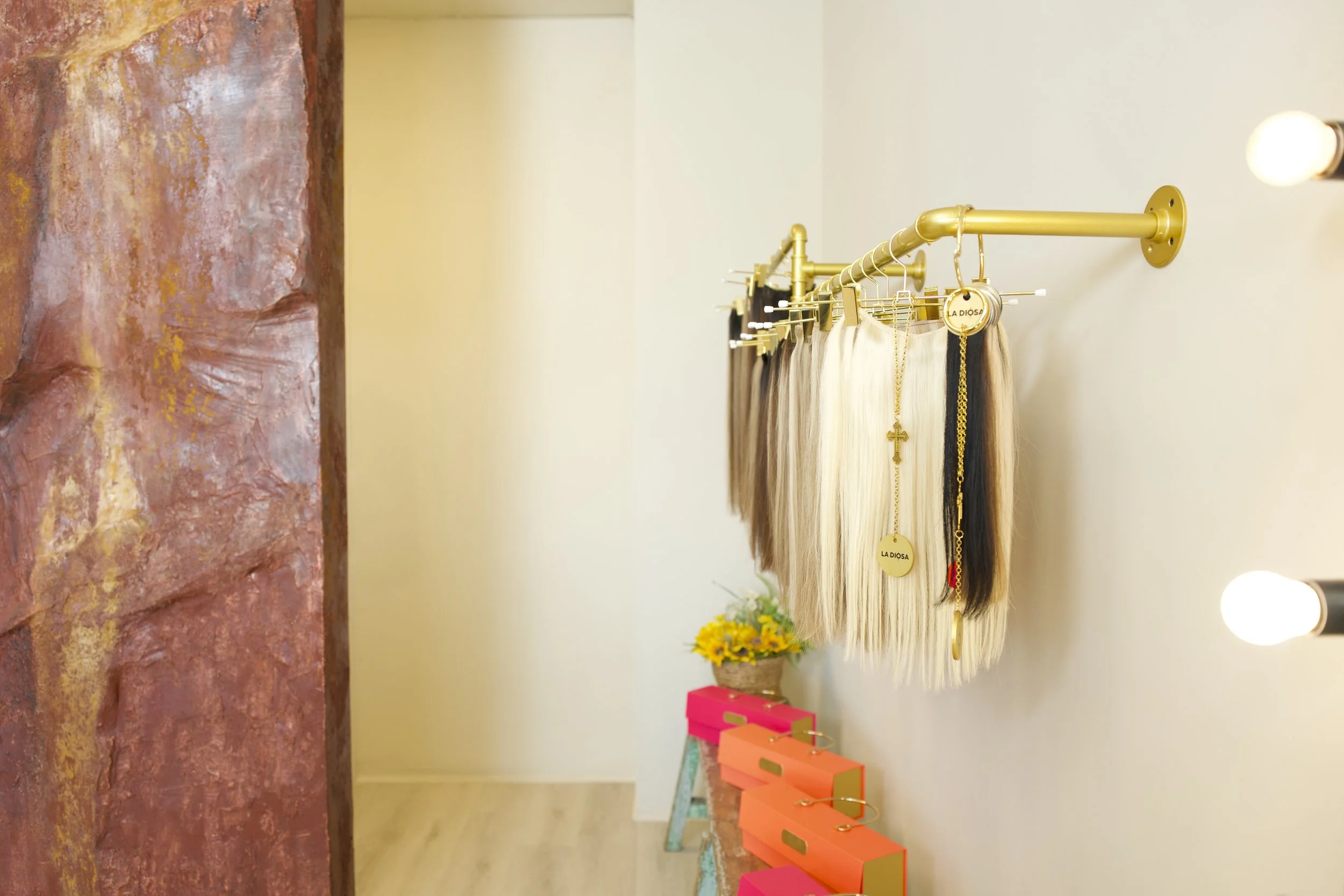 Mostrador con pelucas de cabello largo en tonos rubio, negro y castaño, adornadas con collares dorados y etiquetas que dicen 'La Dosa', en un espacio decorado con una pared rosada y flores amarillas en una maceta, y cajas rosa en una mesa auxiliar.
