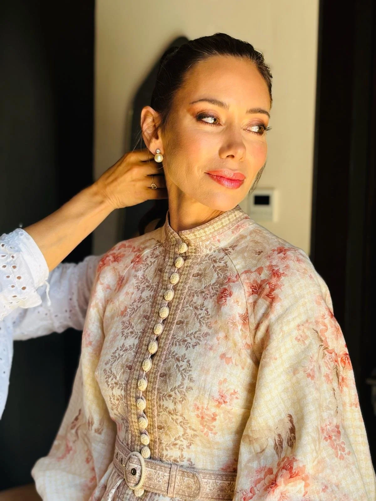 Mujer con peinado recogido, usando un vestido beige con detalles florales y botones en el frente, y pendientes de perlas, siendo ajustada en el cuello por otra persona.