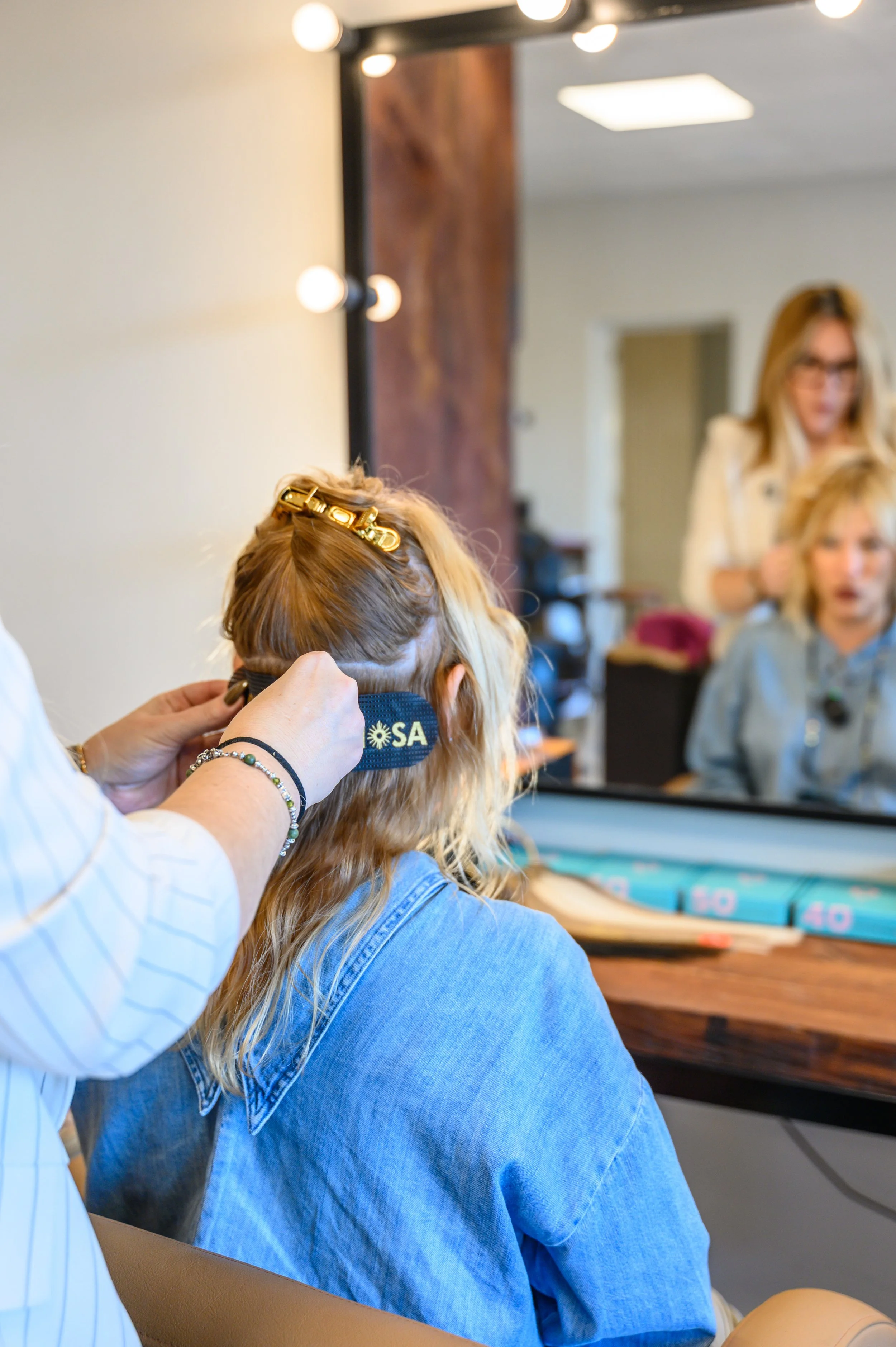 Una persona con cabello rubio y rizado con una pinza dorada, que lleva puesta una pulsera de perlas, se está cepillando el cabello frente a un espejo y le colocan una banda de peluquería con la palabra 'USA'.