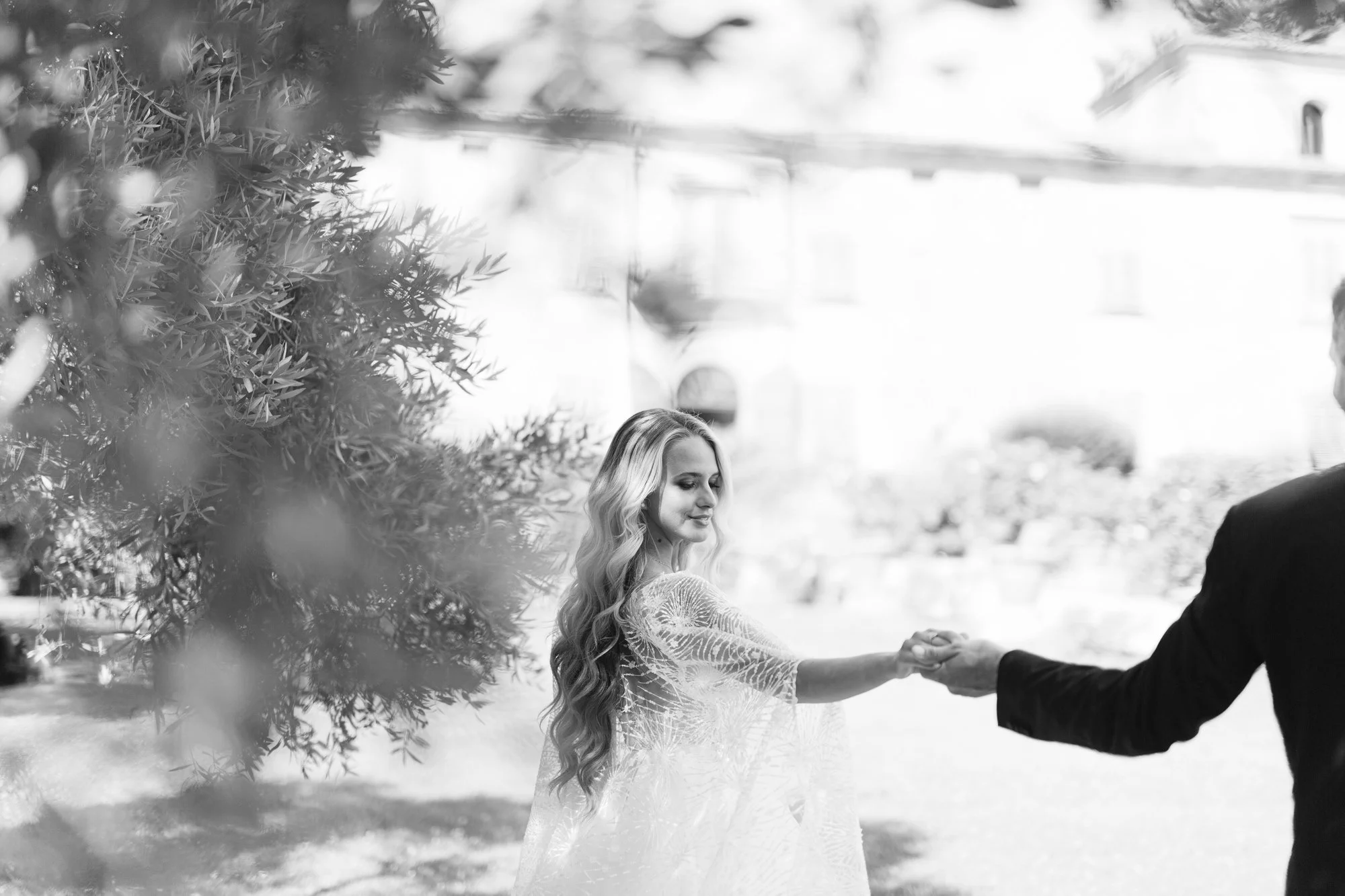 A woman with long wavy hair wearing a lace dress holding hands with a man in a suit, outside with trees and a building in the background, black and white photo.