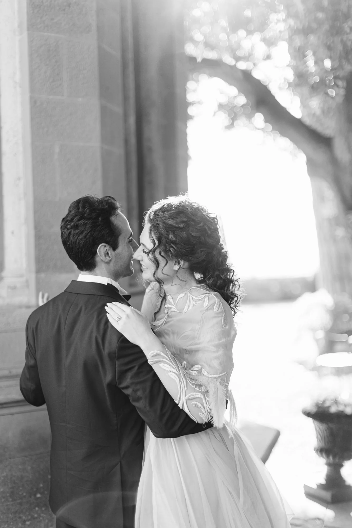 Black and white photo of a bride and groom sharing an intimate moment outdoors near a building, with trees and sunlight in the background.