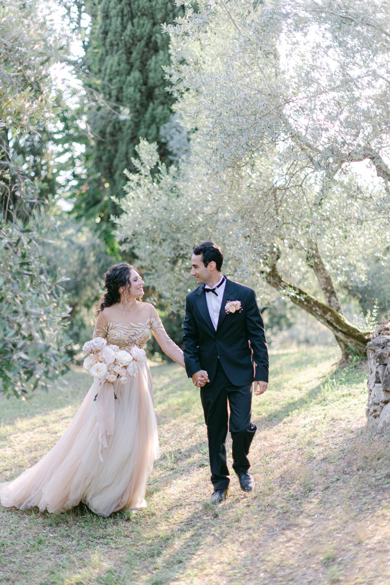 A bride and groom walking outdoors holding hands, surrounded by trees and greenery, during their wedding.