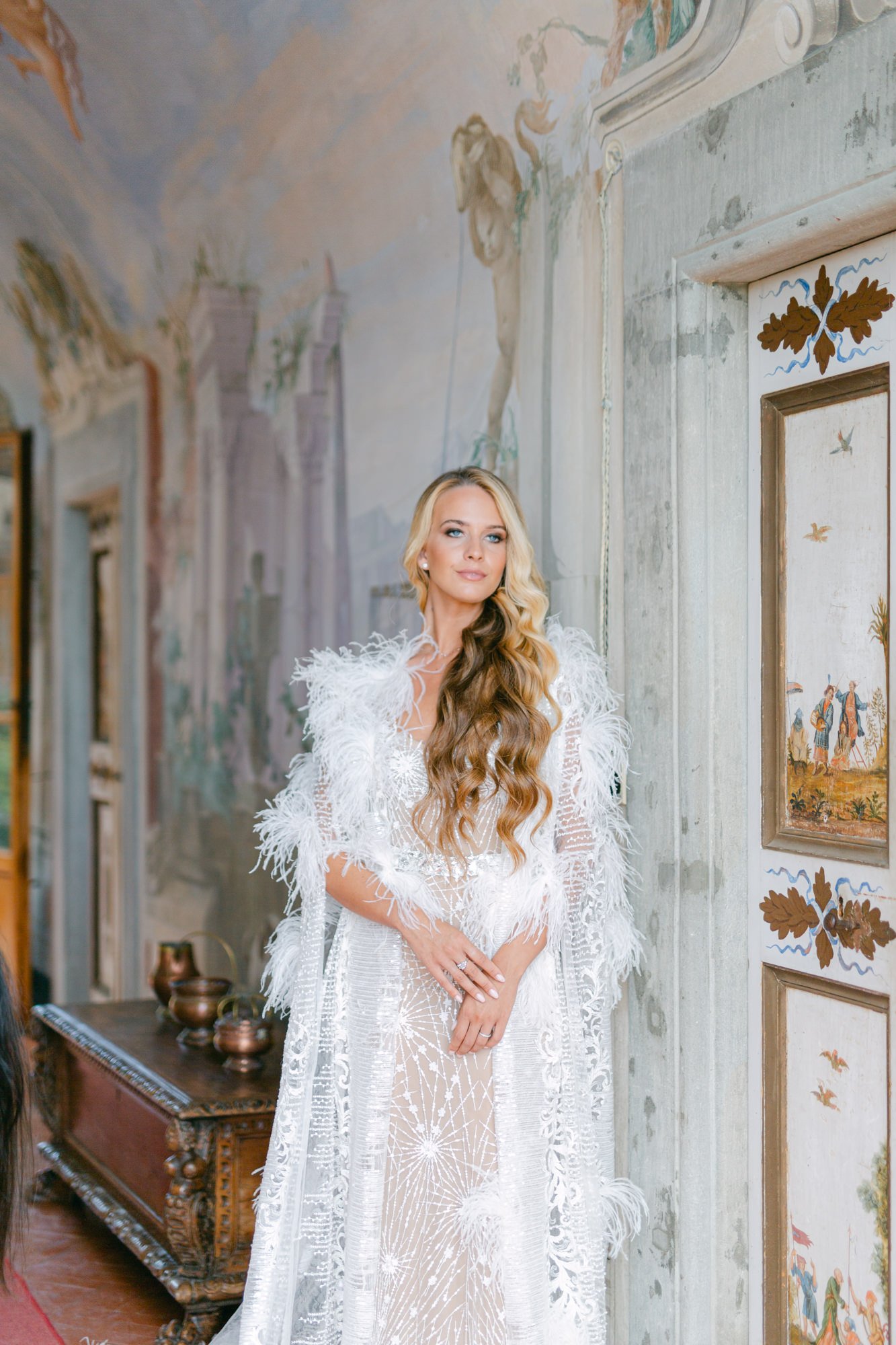 A woman with long, wavy blonde hair wearing a white dress with feather details standing indoors against a painted wall with biblical-style artwork and a decorated door.