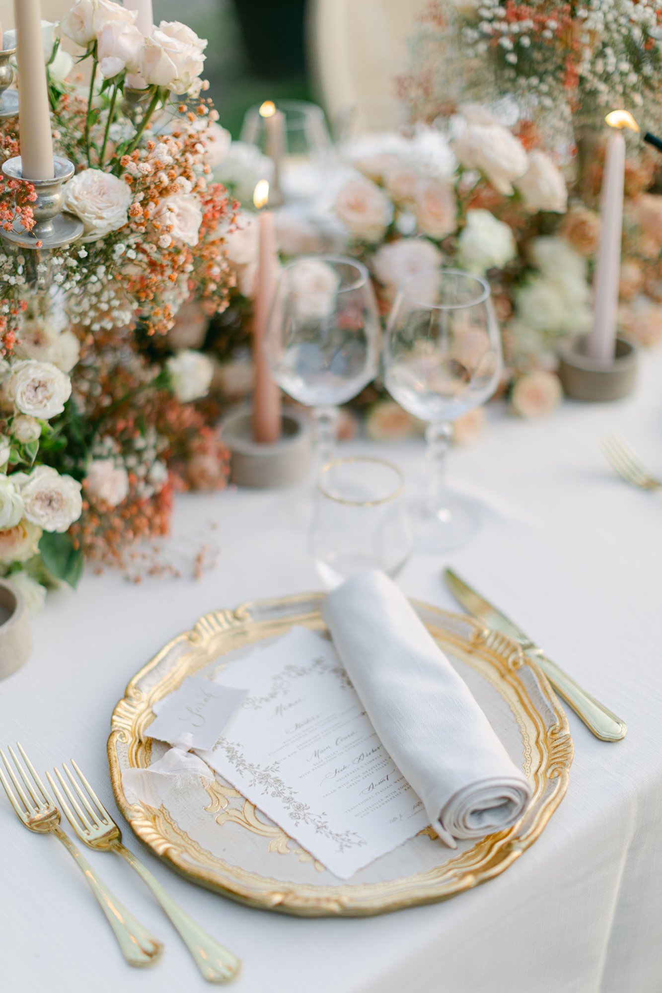 Elegant table setting with a gold-rimmed plate, a rolled white napkin, two wine glasses, a menu card, and floral centerpieces with white and peach roses and baby's breath, accented by tall candles.