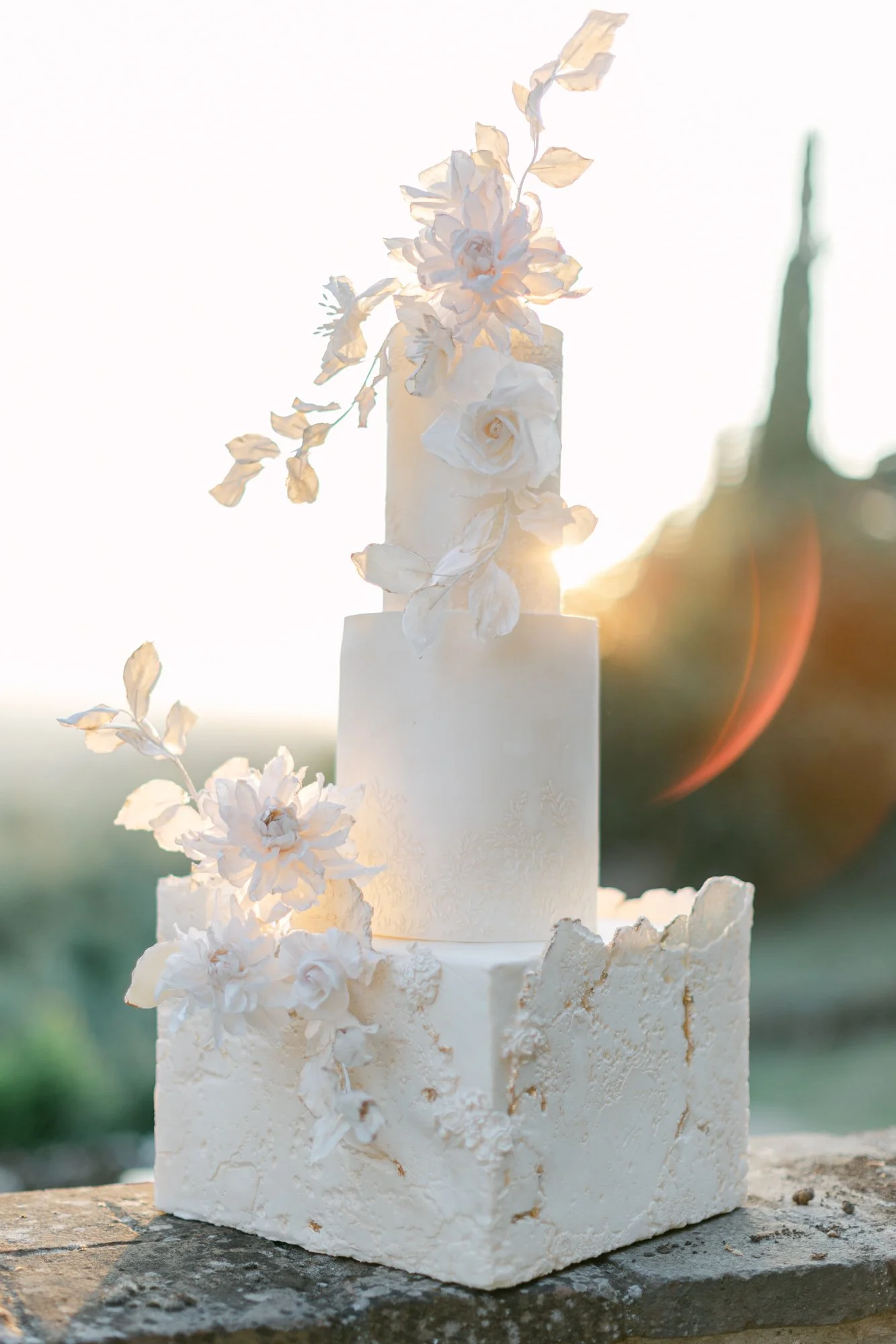 A white tiered wedding cake decorated with white flowers and leaves, set outdoors during sunset.