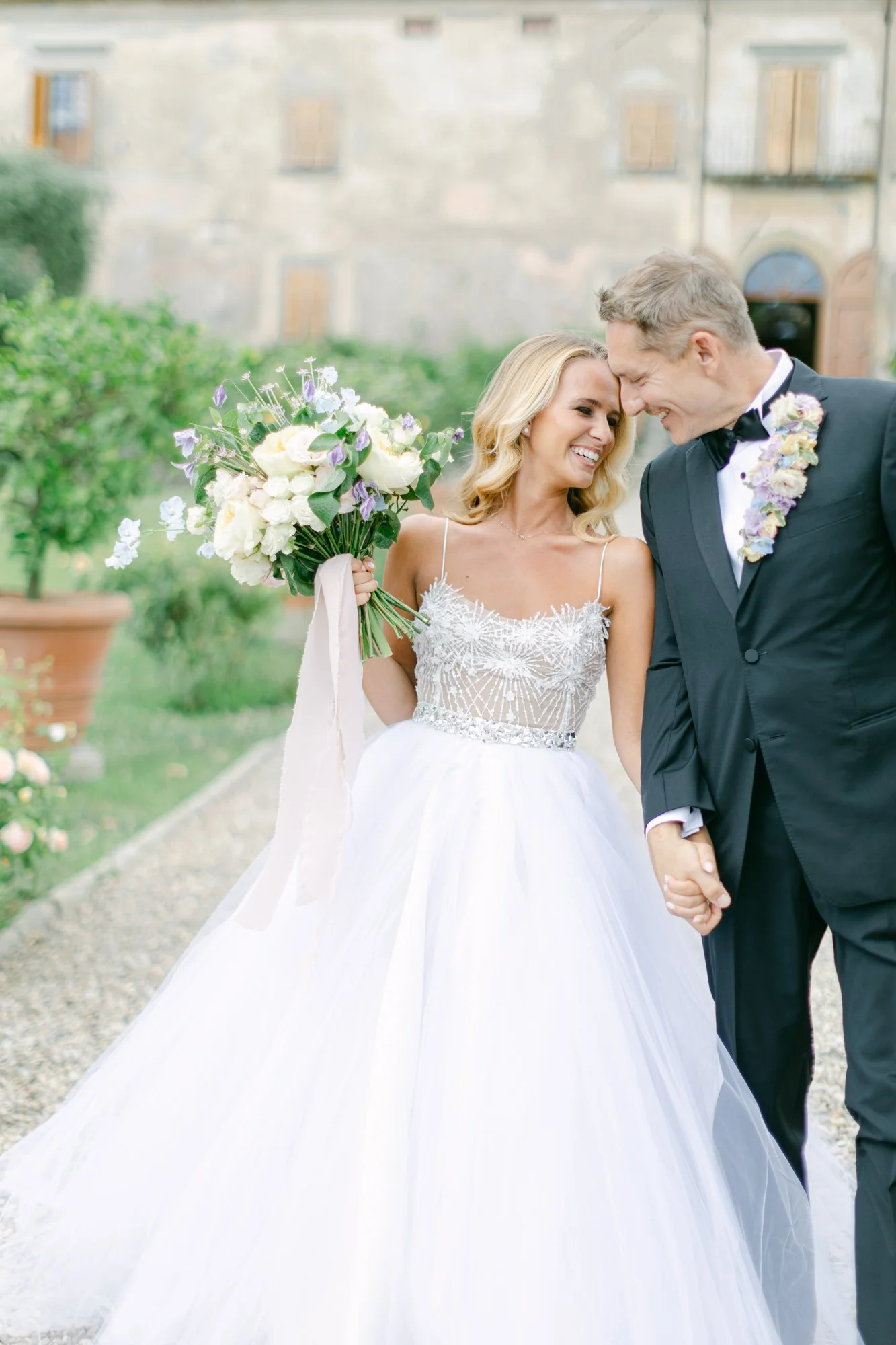 A bride and groom smiling and holding hands outdoors, with the bride holding a bouquet of flowers.