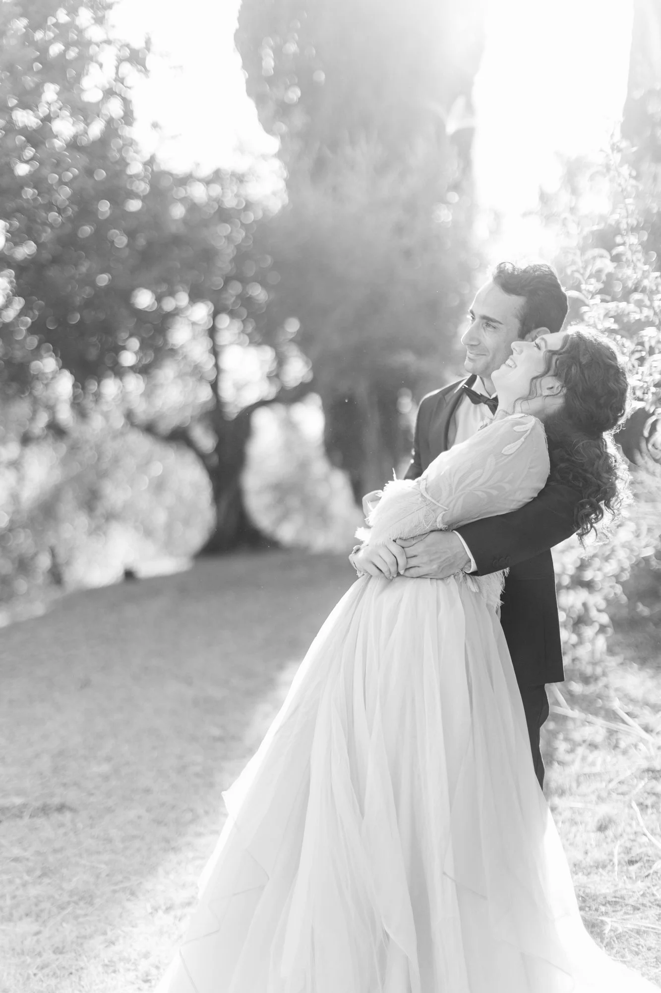 A black and white photo of a joyful couple, with the man in a tuxedo and the woman in a wedding dress, embracing outdoors in a park or garden with trees in the background.