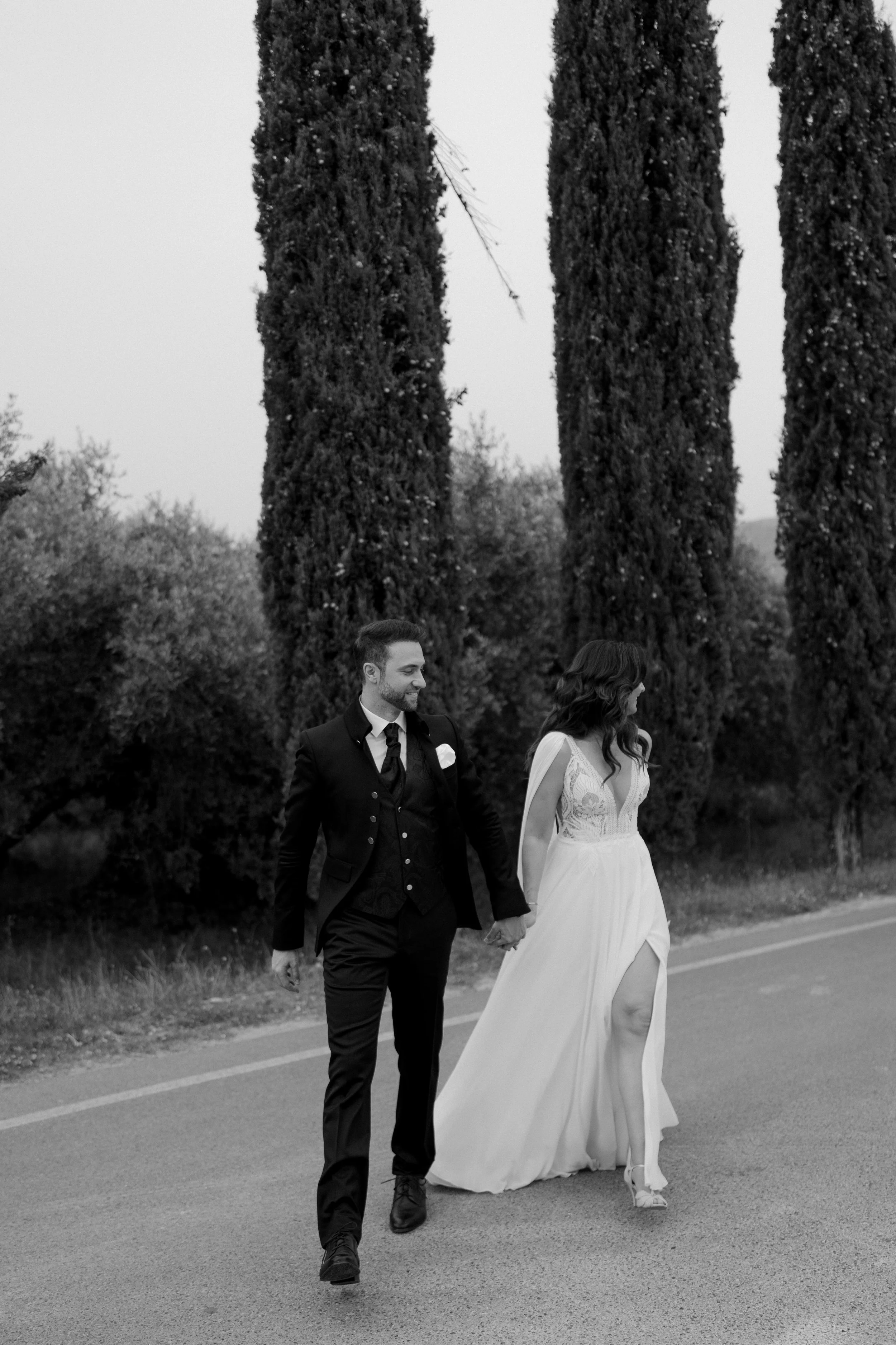 A black-and-white photo of a newlywed couple holding hands walking along a road, with tall trees in the background.