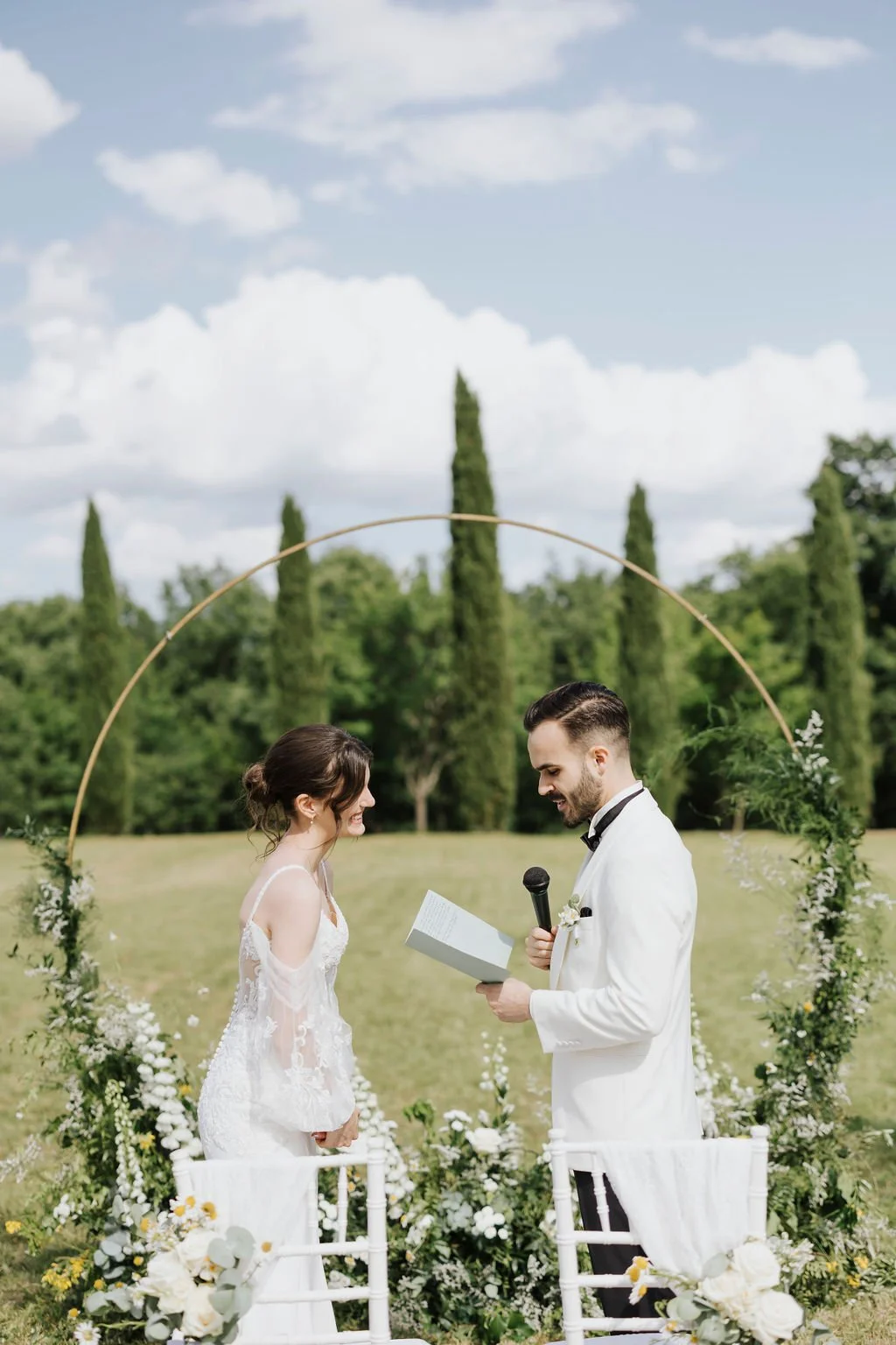 A bride and groom getting married outdoors under a decorative arch, surrounded by flowers, with green trees and a cloudy sky in the background.