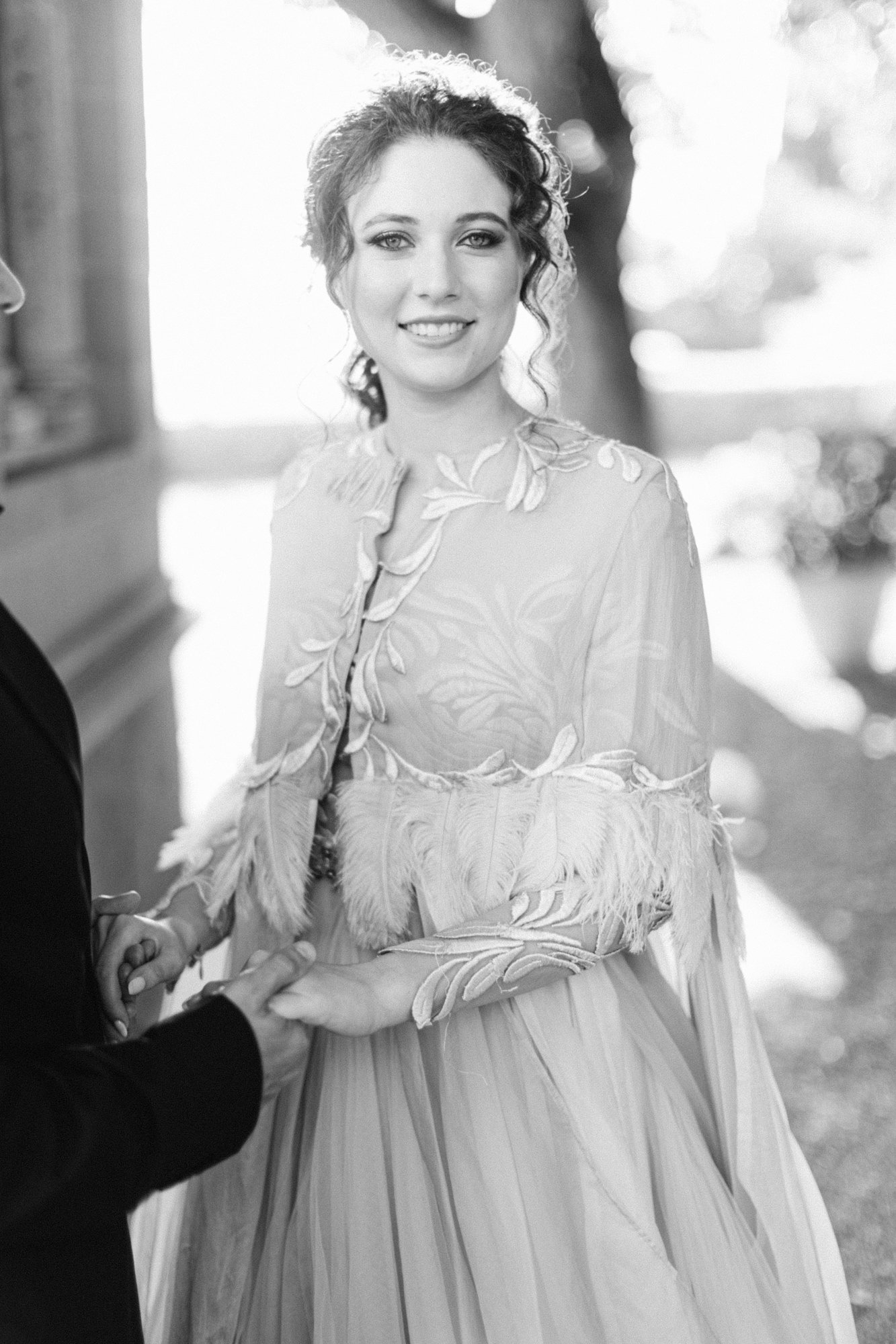 A woman in an elegant dress holding hands with a man during a wedding or formal event. The woman is smiling and has curly hair, with soft sunlight in the background.