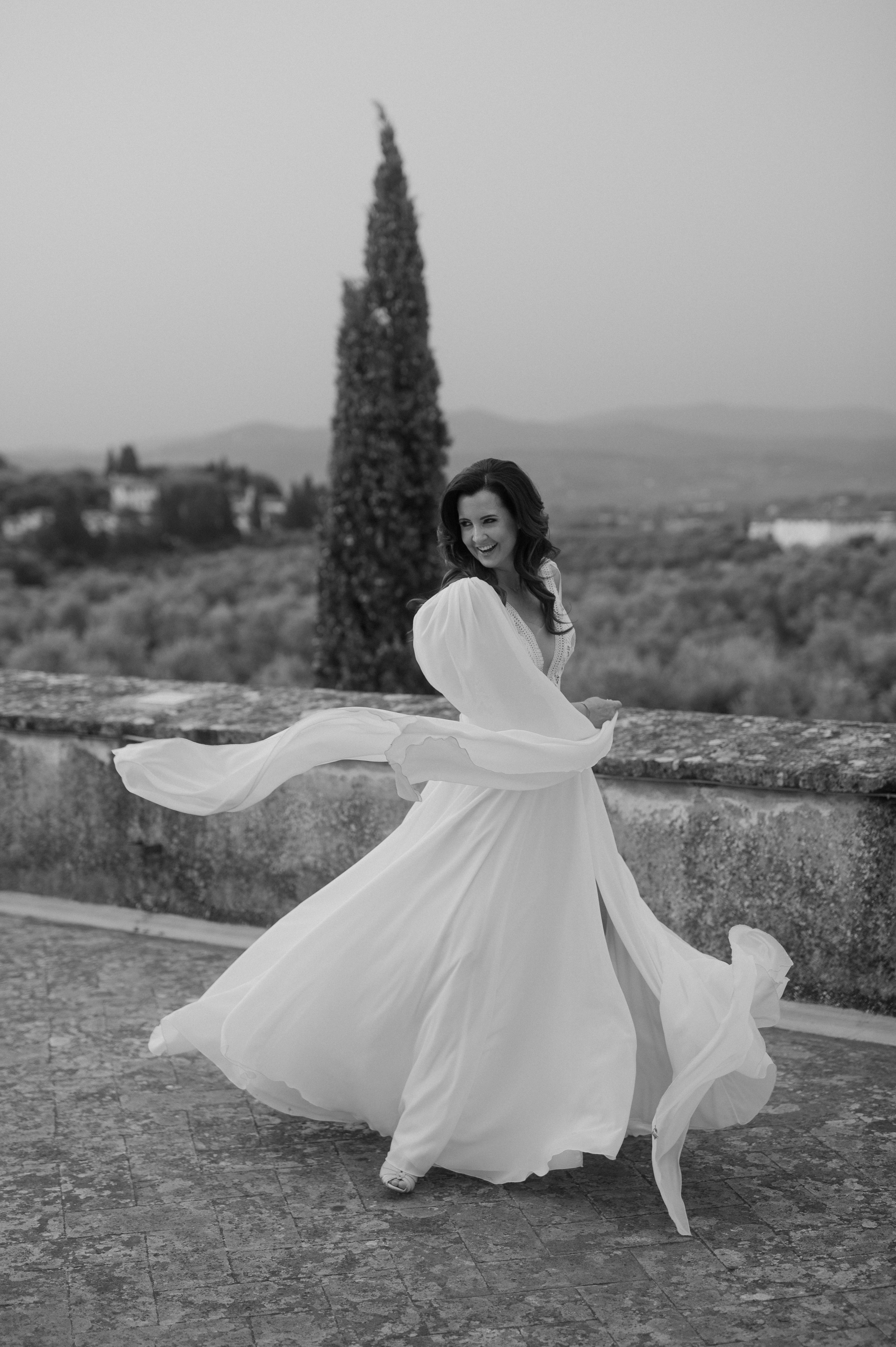 A woman in a long white dress twirls on a stone path outdoors, with a tall cypress tree and distant hills in the background, in black and white.