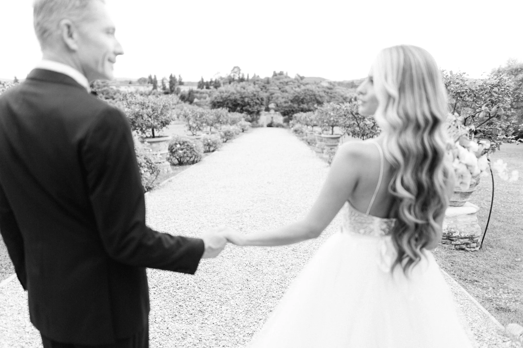A black and white photo of a couple holding hands and facing each other on a garden pathway, with trees and shrubs in the background.