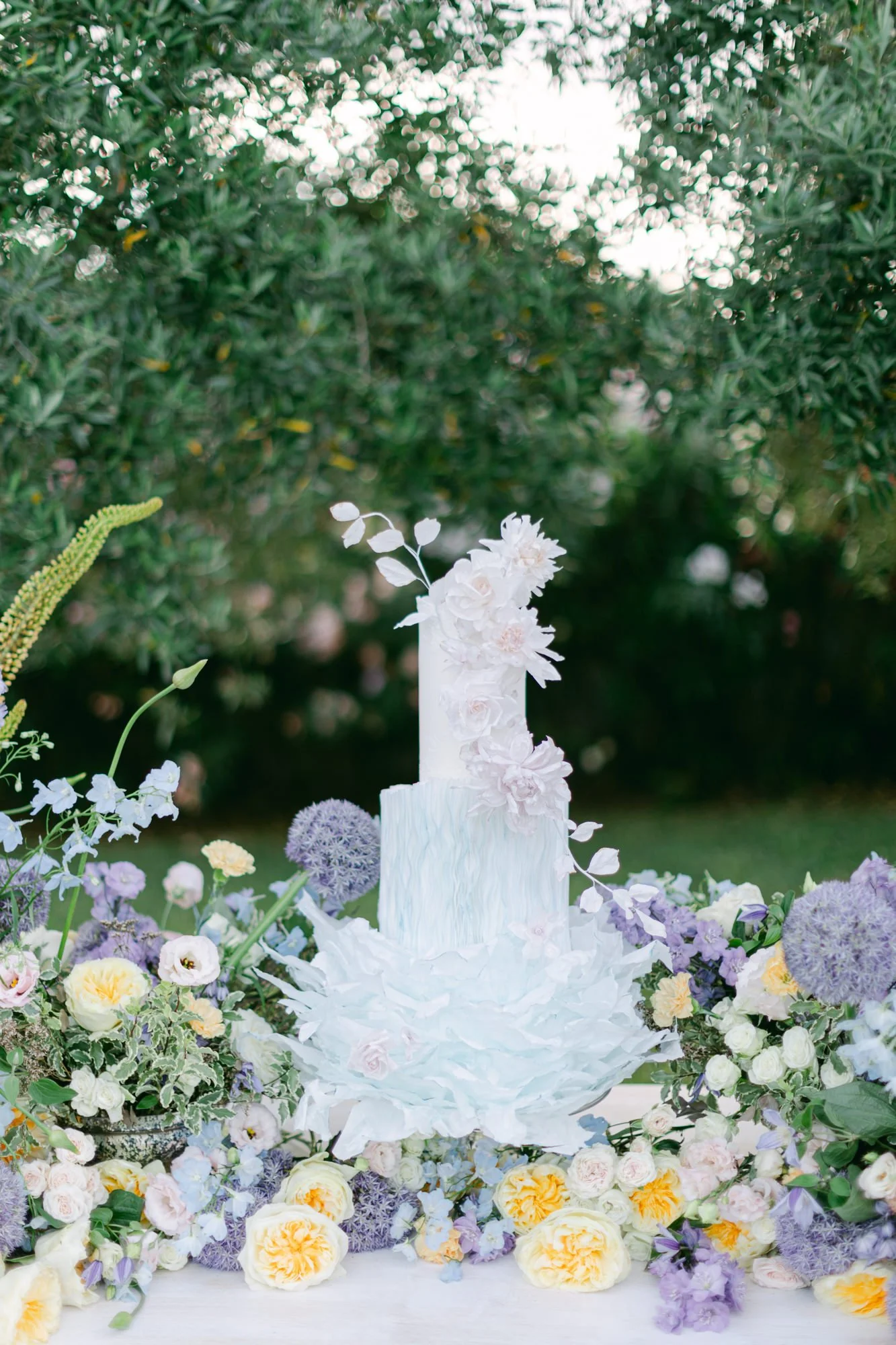 Elegant wedding cake decorated with white and blush flowers on a table surrounded by colorful flowers and greenery.