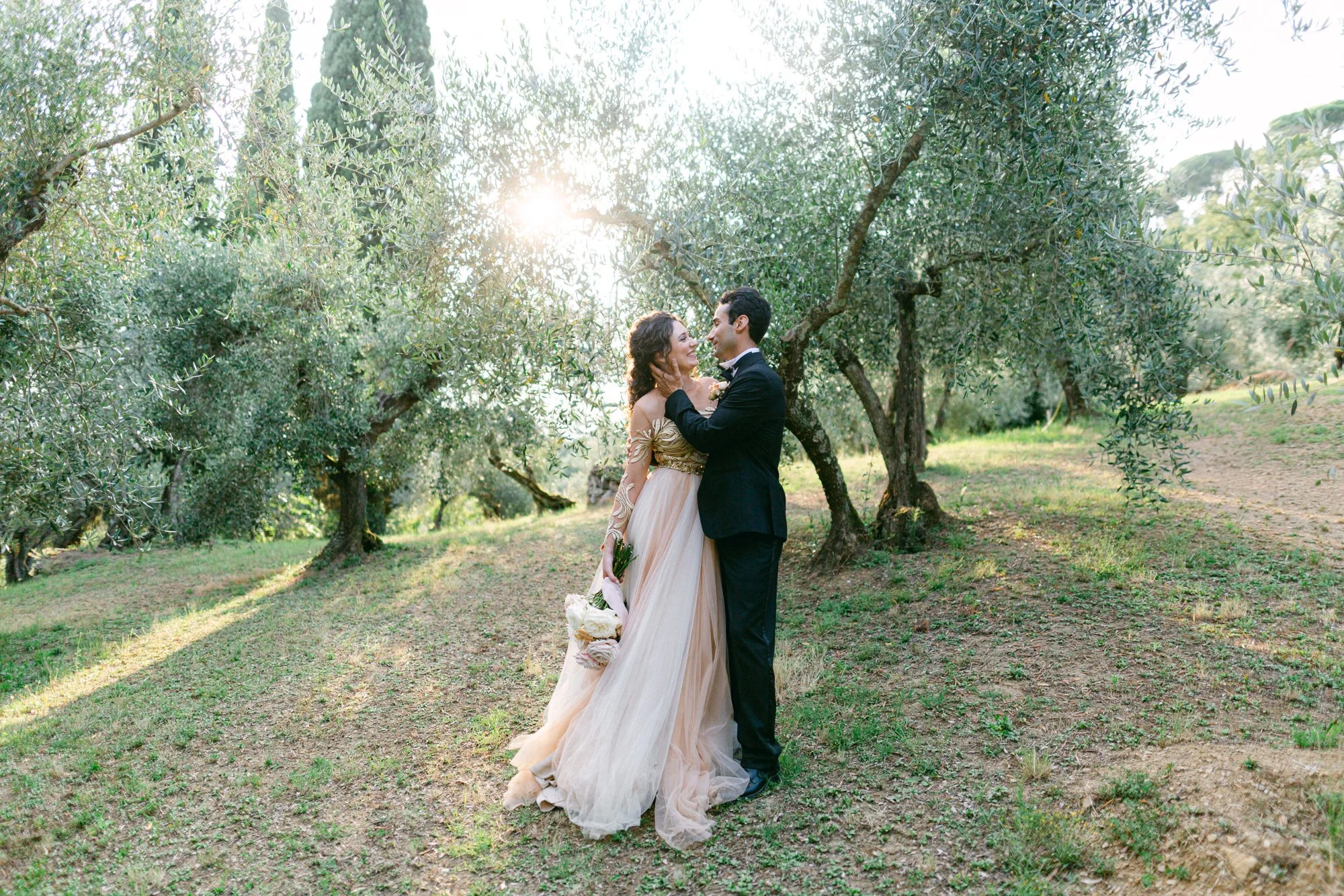 A bride and groom standing close together in an olive orchard during sunset, looking at each other lovingly, with the bride holding a bouquet of white flowers.