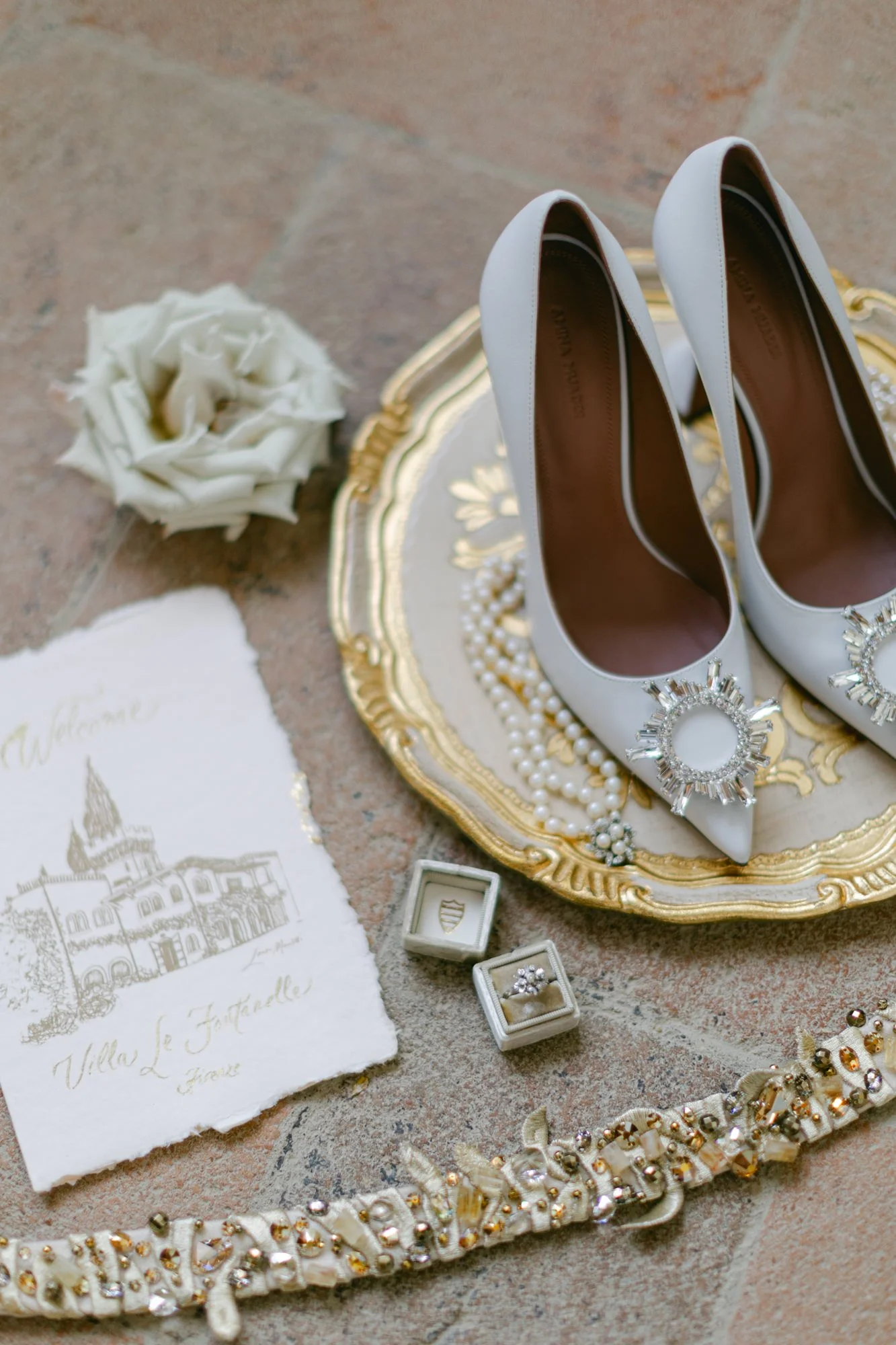 White high-heeled shoes with jeweled embellishments on the front, placed on a decorative gold and white tray, surrounded by jewelry, a white flower, a handwritten note, and two rings in small boxes.