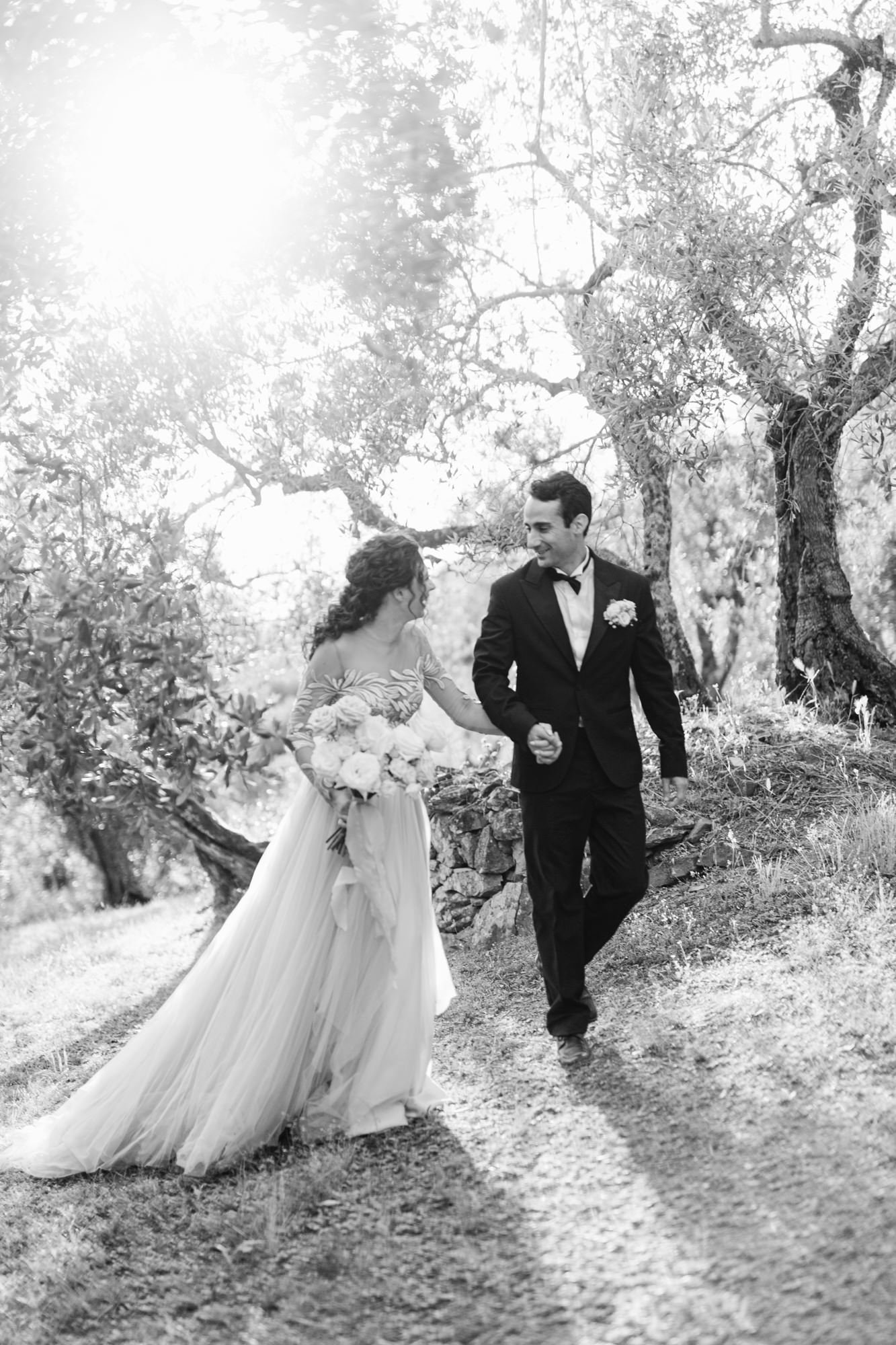 Black and white photograph of a bride and groom walking hand in hand outdoors, surrounded by trees and sunlight filtering through the branches. The bride is carrying a bouquet and wearing a wedding dress, while the groom is in a tuxedo.
