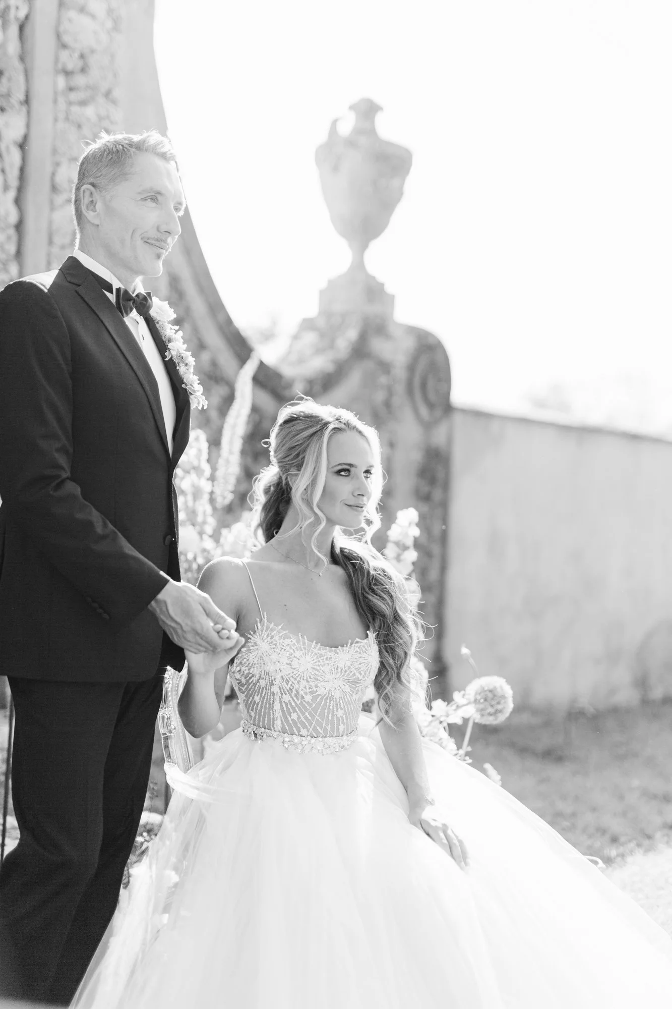Black and white photo of a bride and groom holding hands during their wedding ceremony outdoors, with a stone wall and decorative urns in the background.