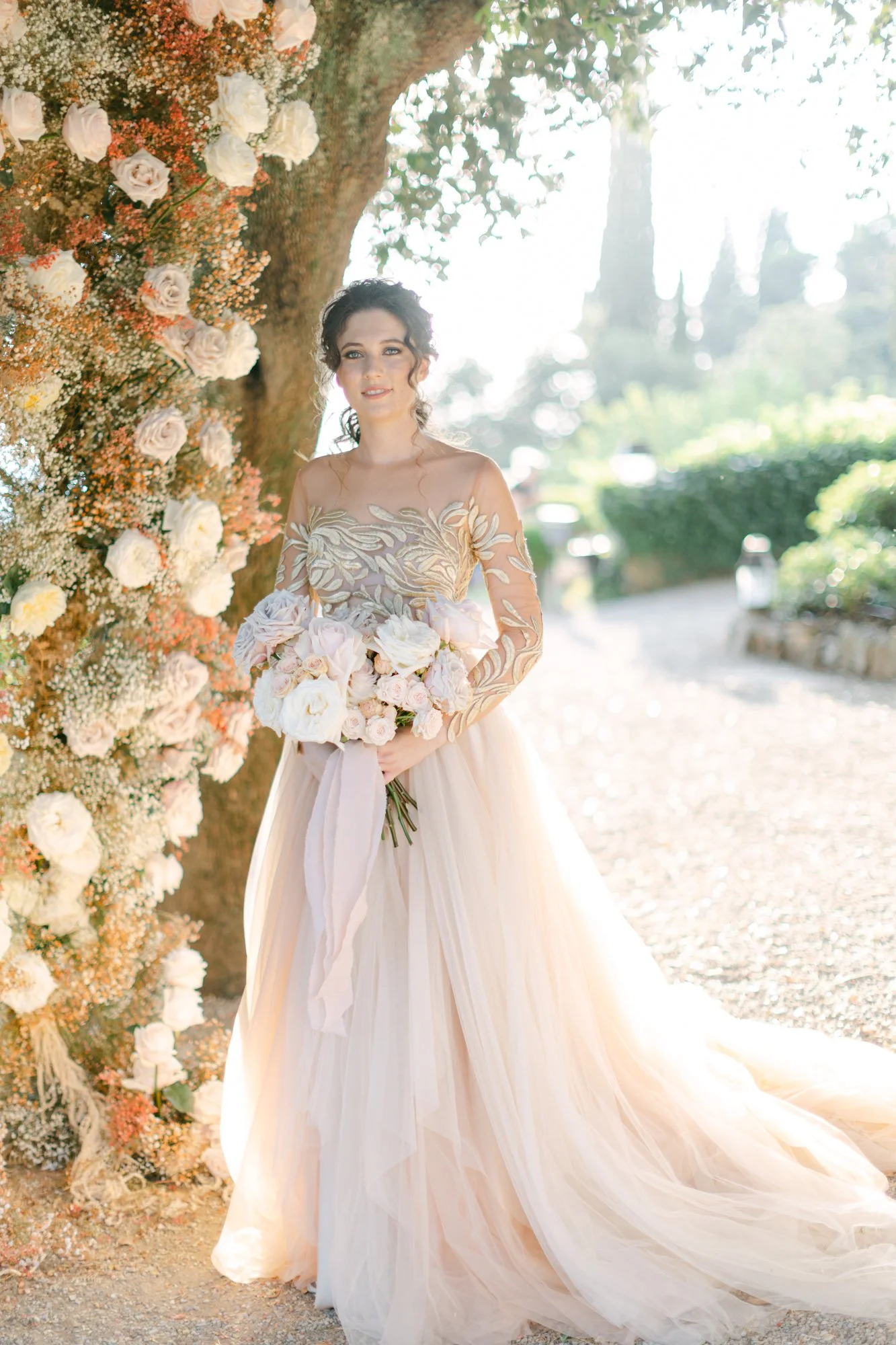 A bride holding a large bouquet of pink and white flowers in front of a floral arch with white roses and peach-colored blooms, standing outdoors on a sunny day.