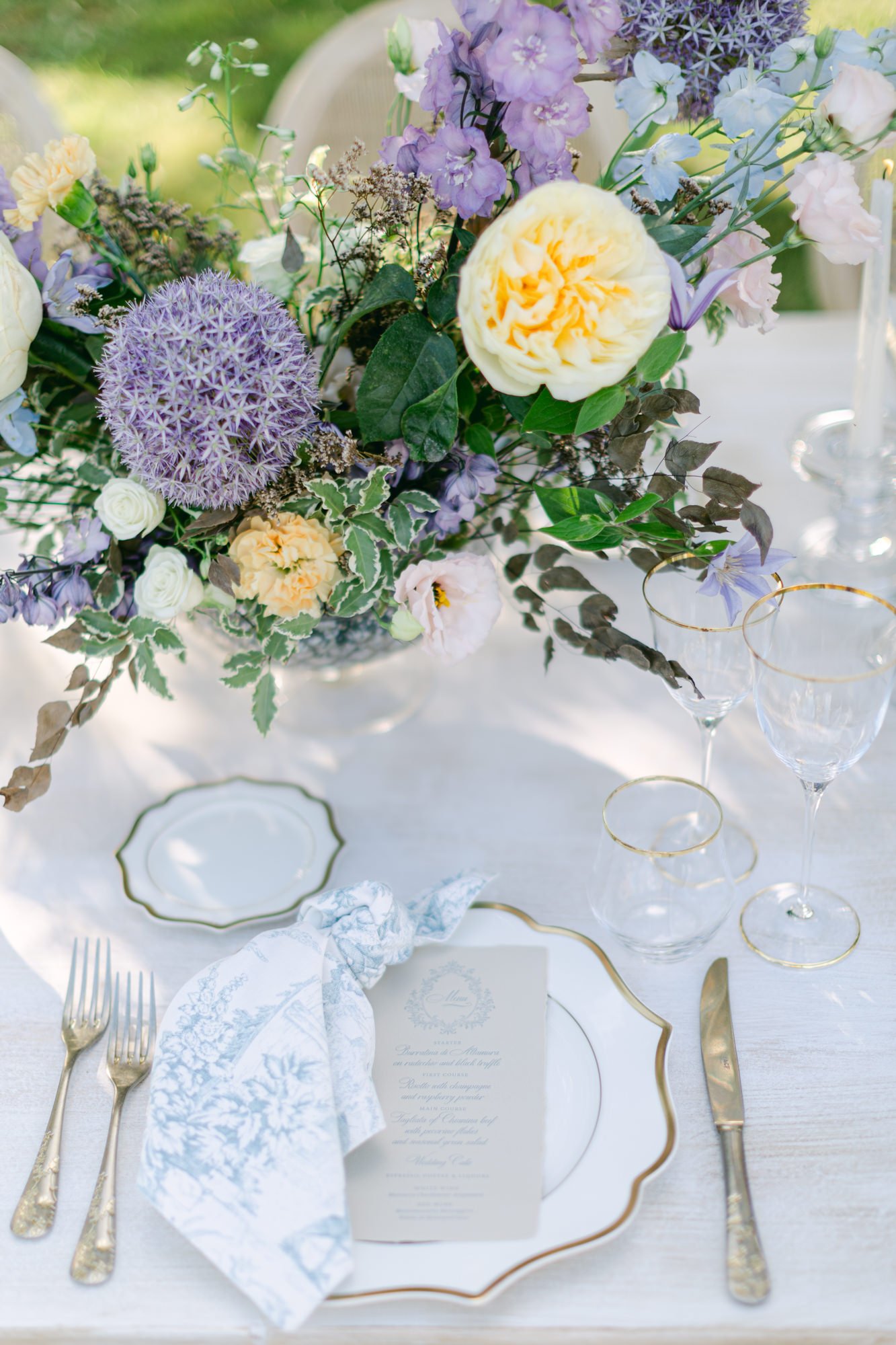 Elegant table setting with a large floral centerpiece featuring purple, white, and cream flowers, fine china with gold trim, a printed menu, a white and blue patterned napkin, and gold-rimmed glassware.
