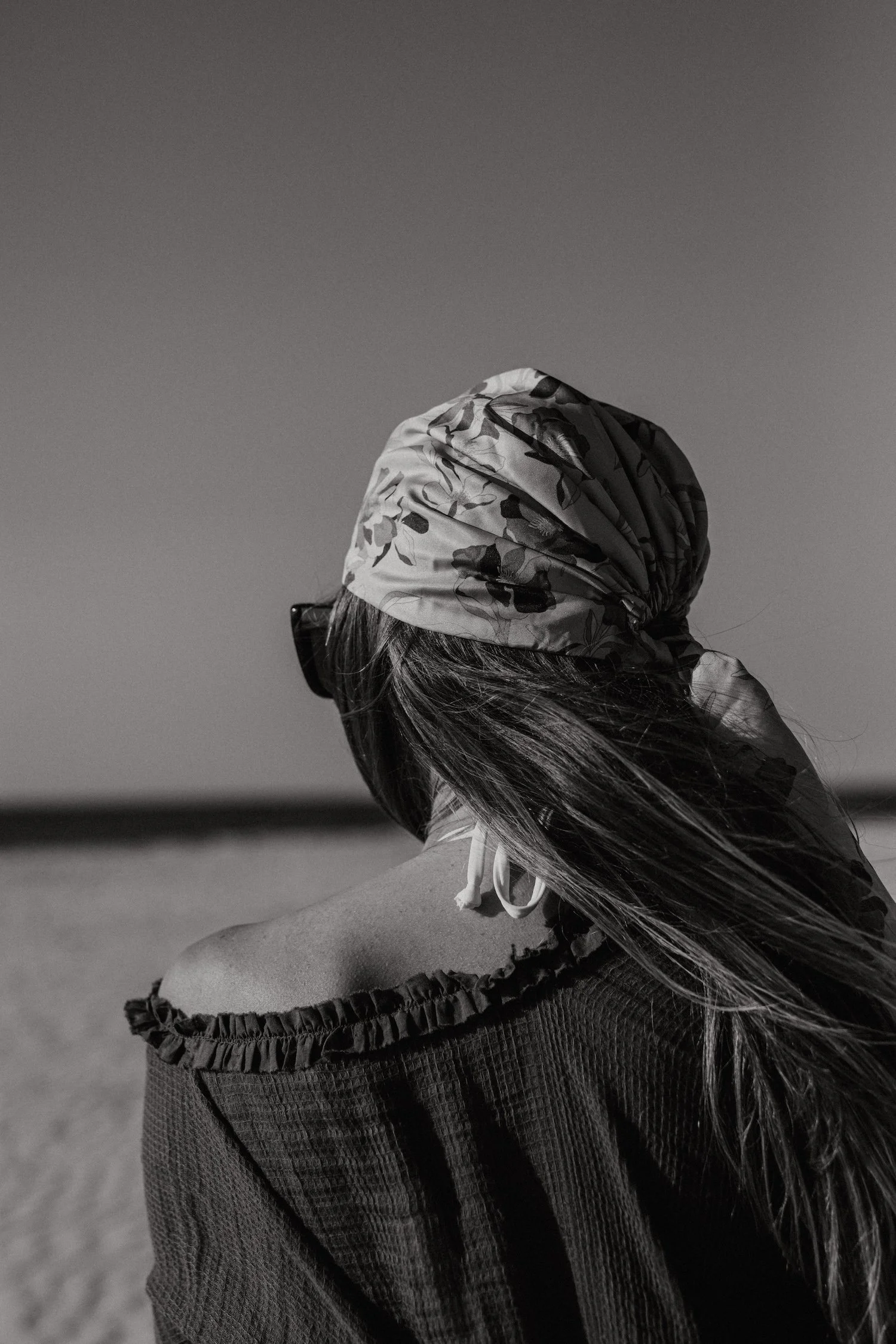 A woman facing away, wearing a headscarf, sunglasses, and an off-the-shoulder top, at the beach in black and white.