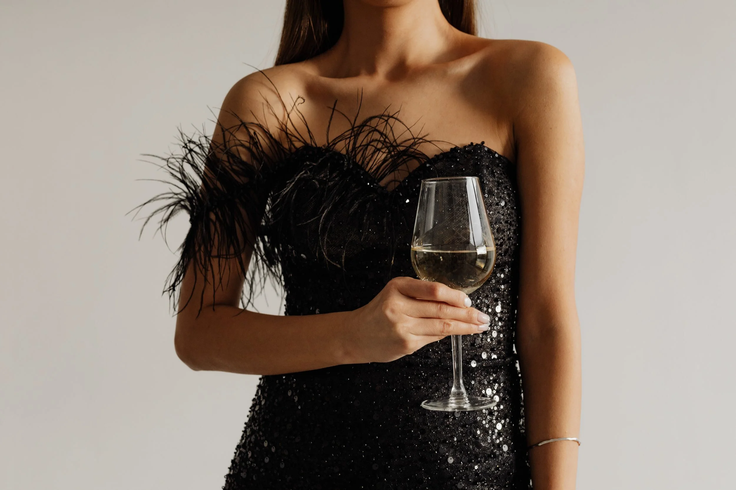 Woman in a black, sequined dress with feather details on the shoulder, holding a glass of white wine.