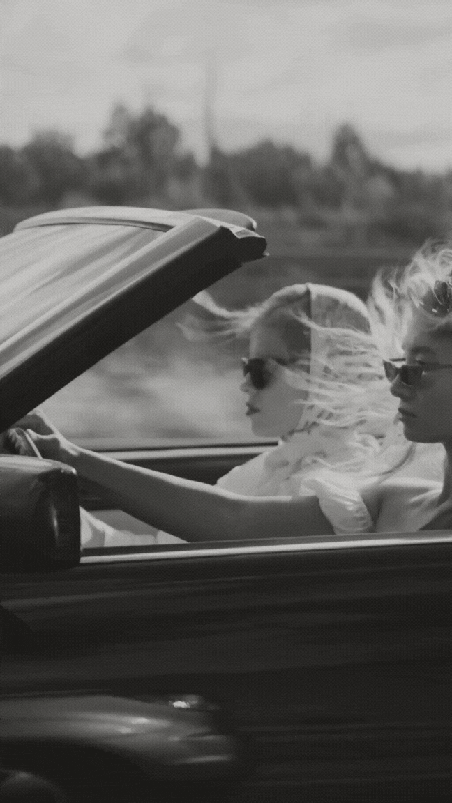 Two women with sunglasses driving a convertible car on the highway, wind blowing their hair, in black and white photo.