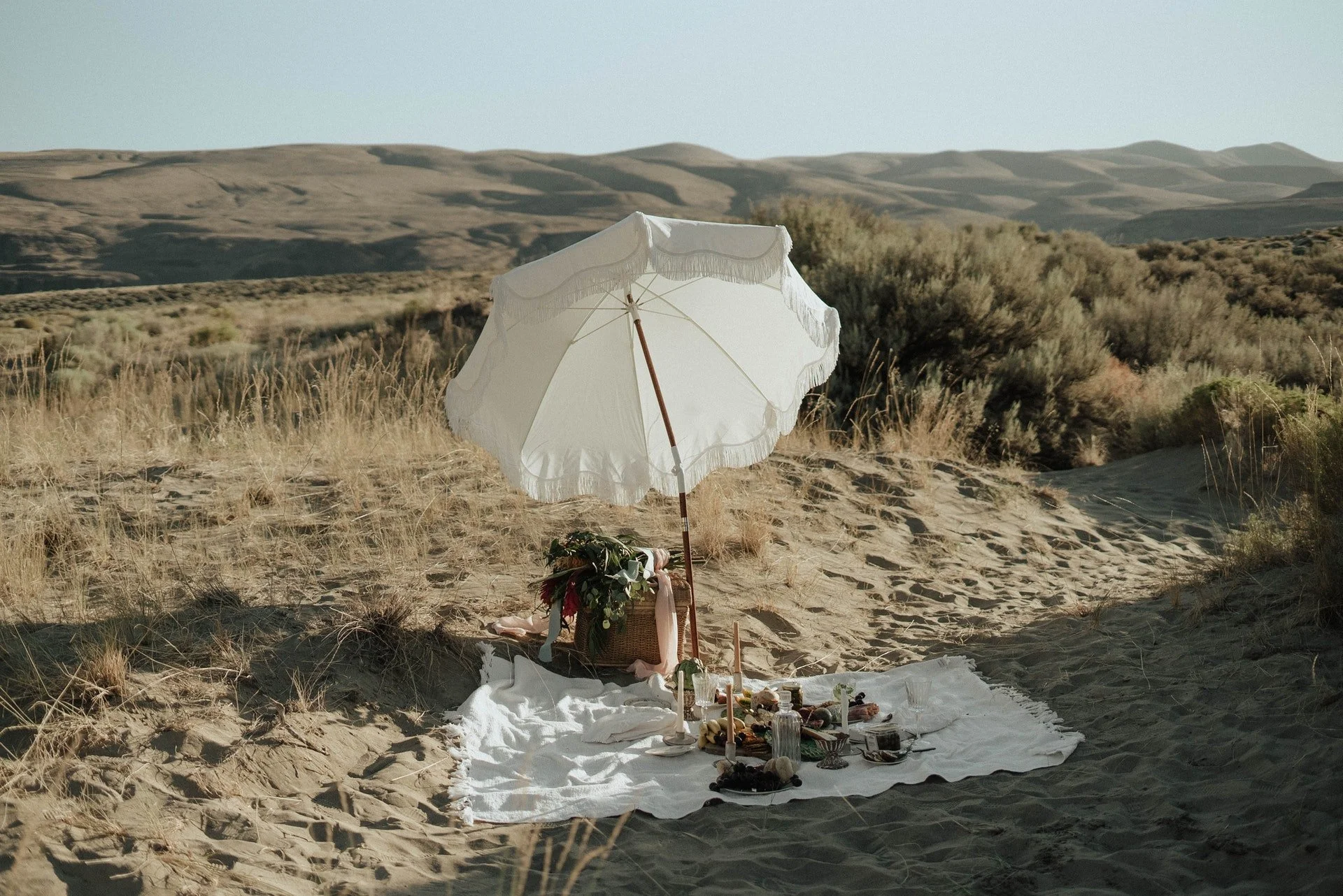 A picnic setup on a white cloth spread on sandy dunes, with a white umbrella providing shade, a basket with flowers, and various food and drink items, in a desert landscape with hills and sparse bushes in the background.