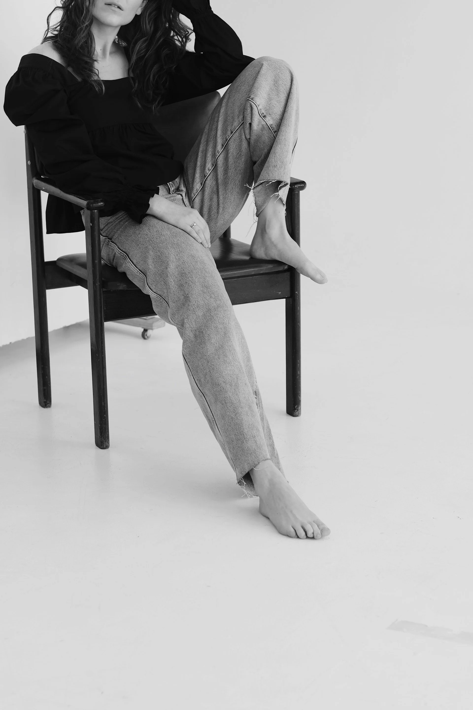 A woman sitting on a wooden chair with one leg crossed over the other, barefoot, holding her head with one hand, dressed in a black top and ripped jeans, against a plain white background.