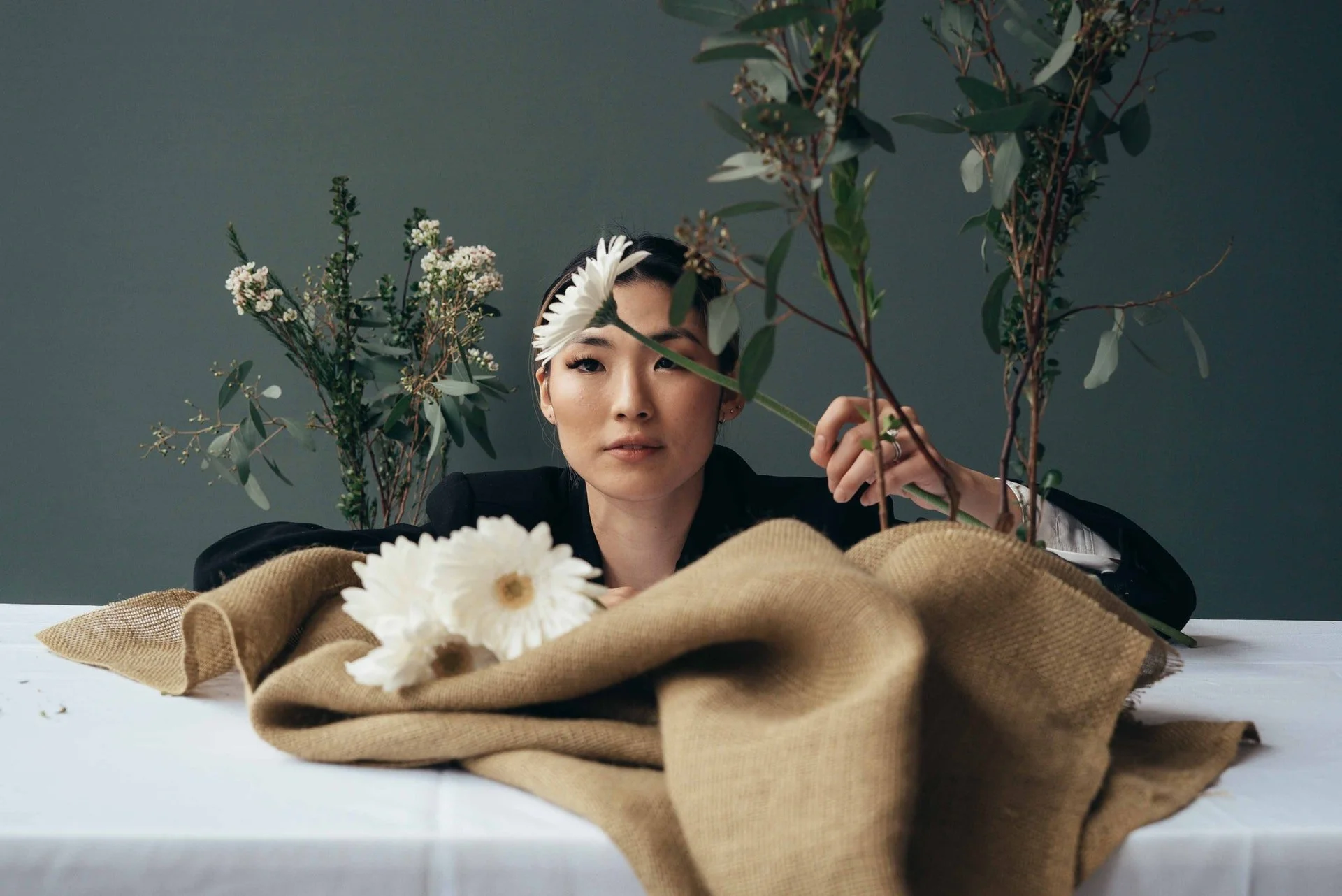 A woman in a black outfit leaning on a table surrounded by flowers and green foliage, with a large white flower headband on her head and white daisies on the table, in front of a plain gray background.