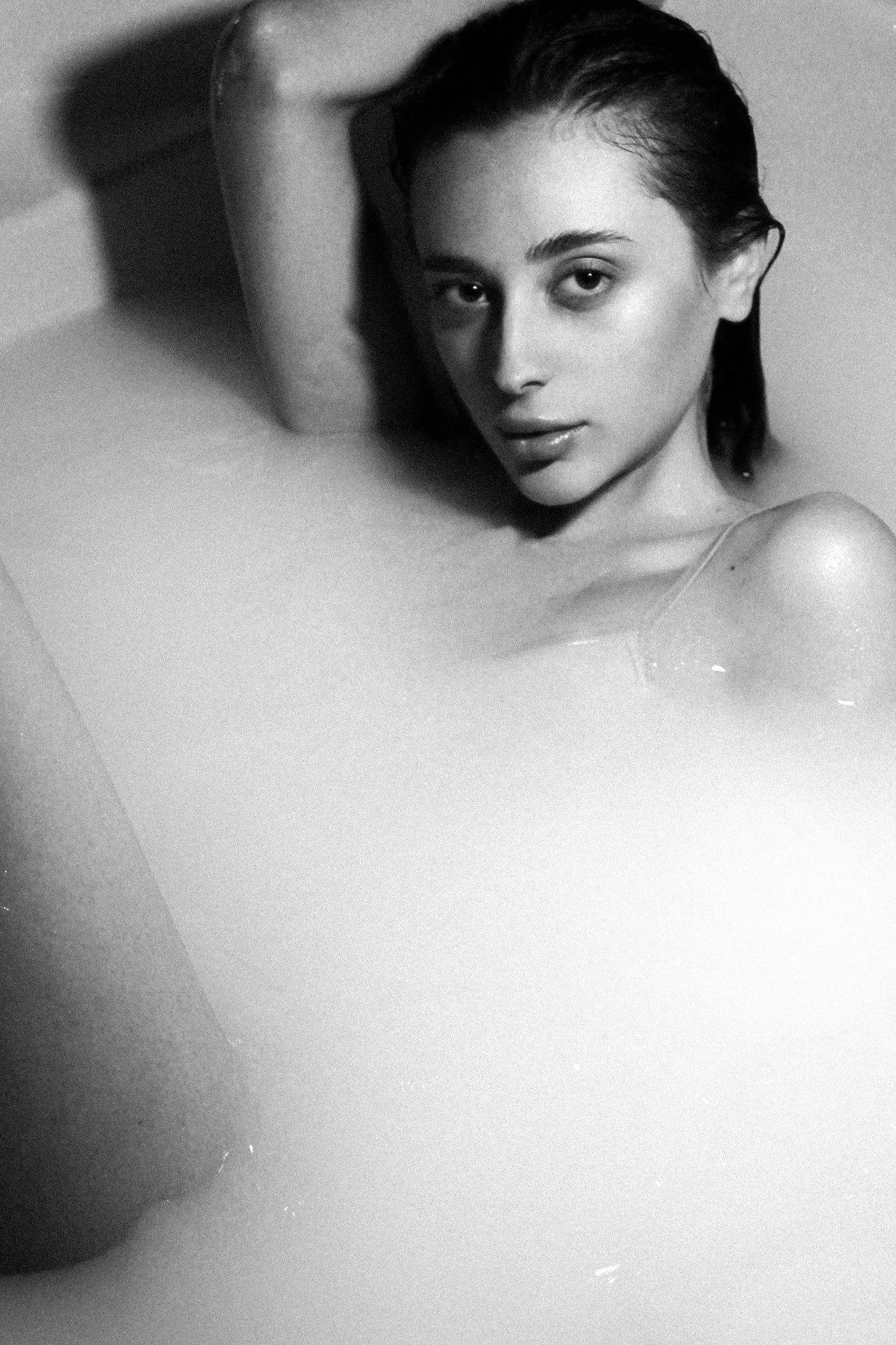 A young woman with wet hair and a relaxed expression lying in a bathtub filled with milky water.