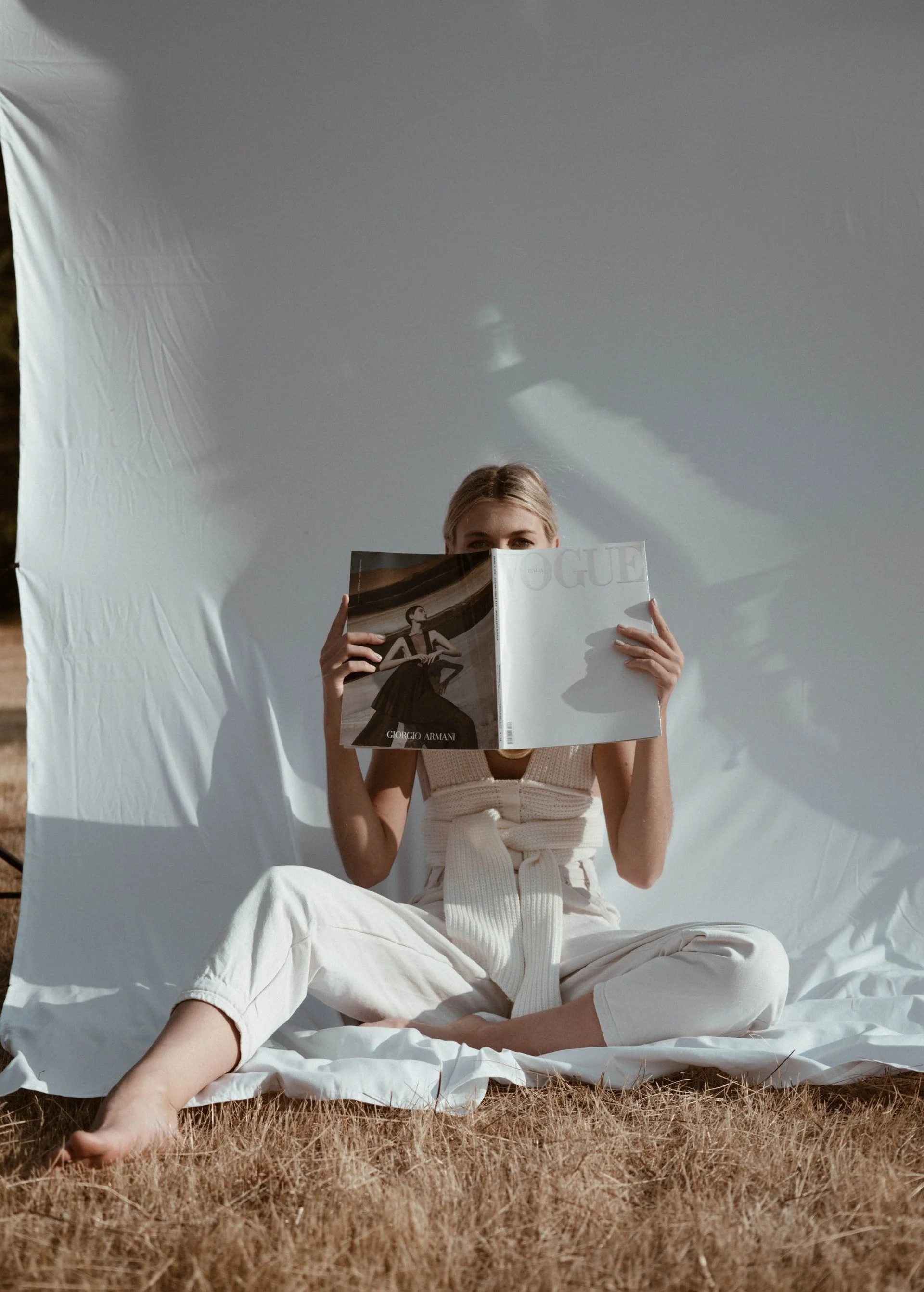A woman sitting outdoors on grass, reading a Vogue magazine, with a large white backdrop behind her.