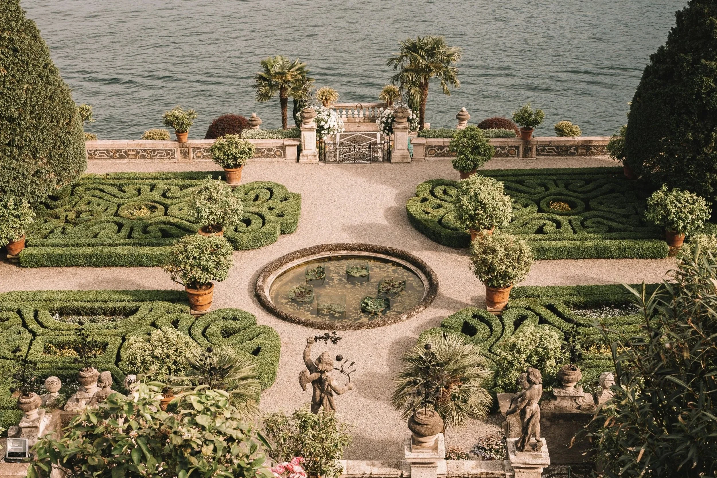 A manicured garden with symmetrical hedges, potted plants, statues, and a fountain, overlooking a body of water in the background.