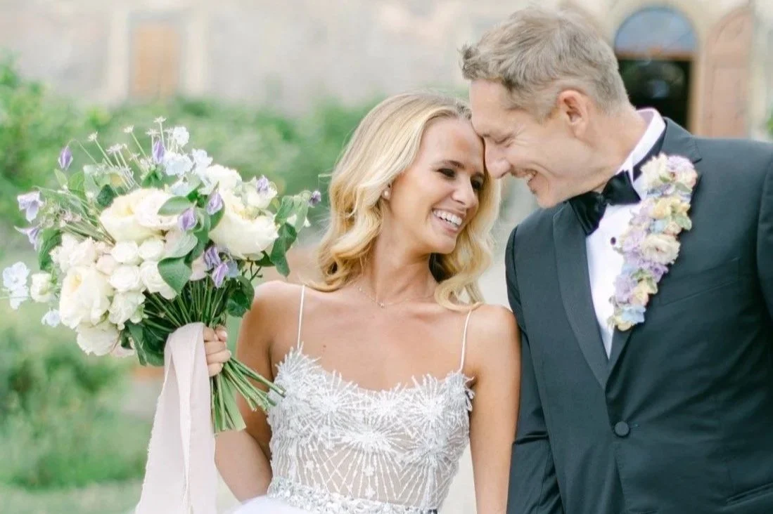 A bride and groom share a joyful moment at their wedding, standing close together with foreheads touching. The bride holds a large bouquet of white and purple flowers and is wearing a white lace wedding dress. The groom is dressed in a black tuxedo with a black bowtie and a floral boutonniere, both smiling happily.
