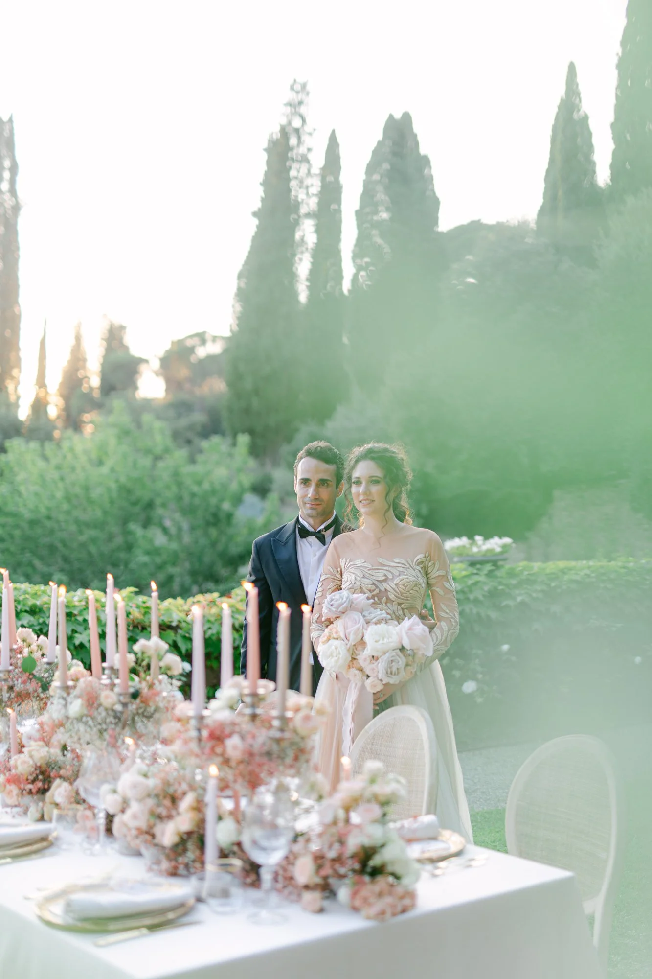 Bride and groom standing outdoors at wedding reception with decorated table, candles, and floral arrangements, with tall cypress trees in the background at sunset.