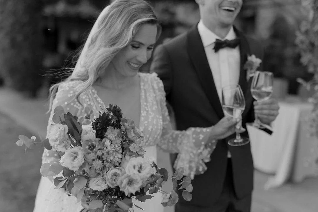 A bride and groom smiling, holding glasses of wine, during their wedding celebration.