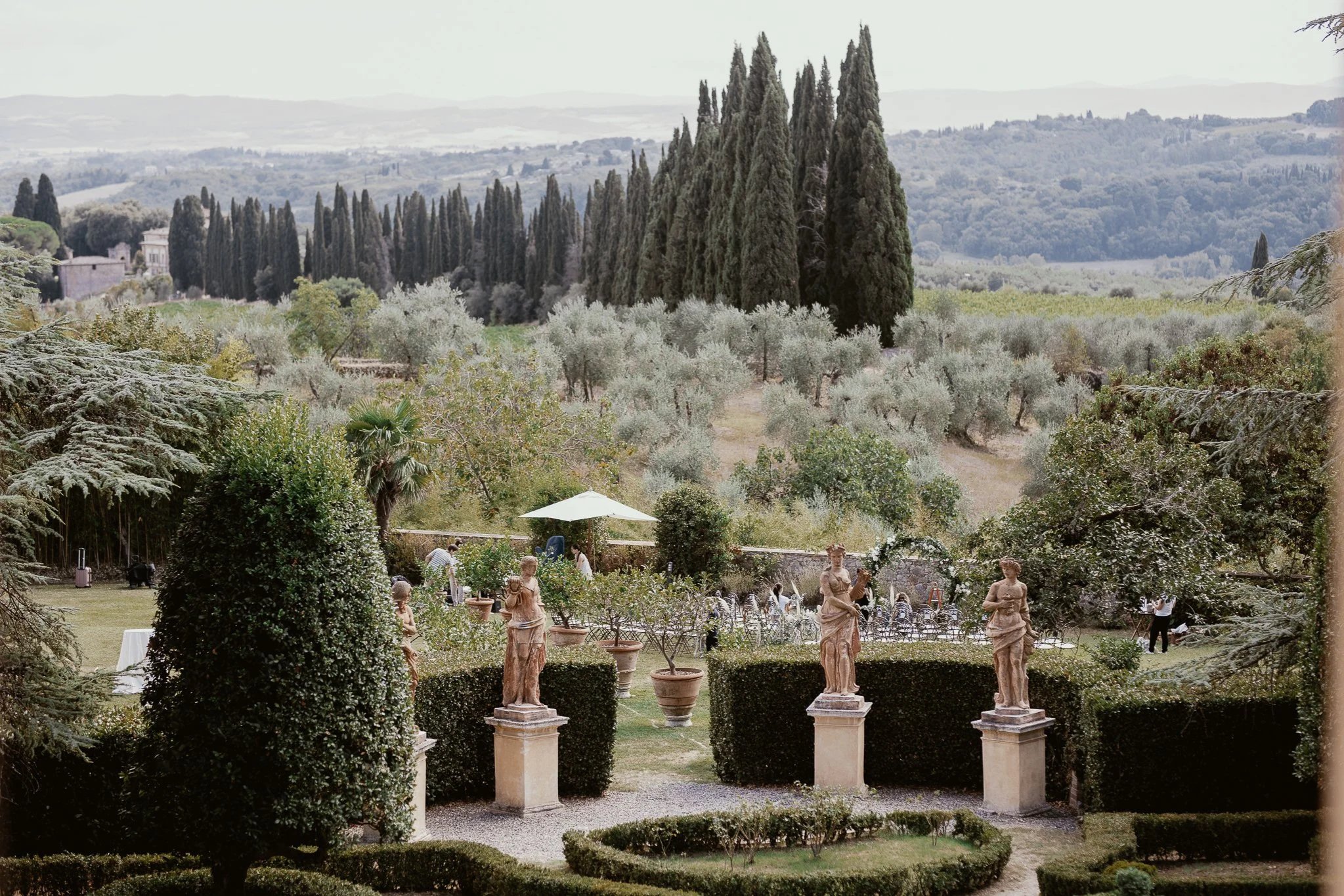 A beautiful outdoor garden scene with classical sculptures, lush greenery, and tall cypress trees in the background.