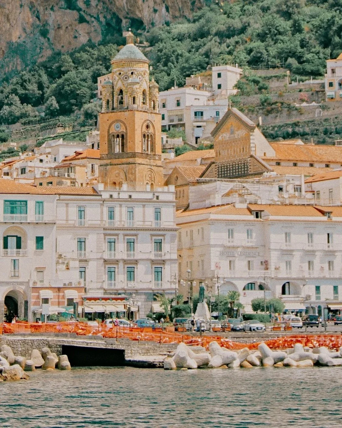 A coastal cityscape with a church tower, surrounding buildings, and a waterfront view at the shore.